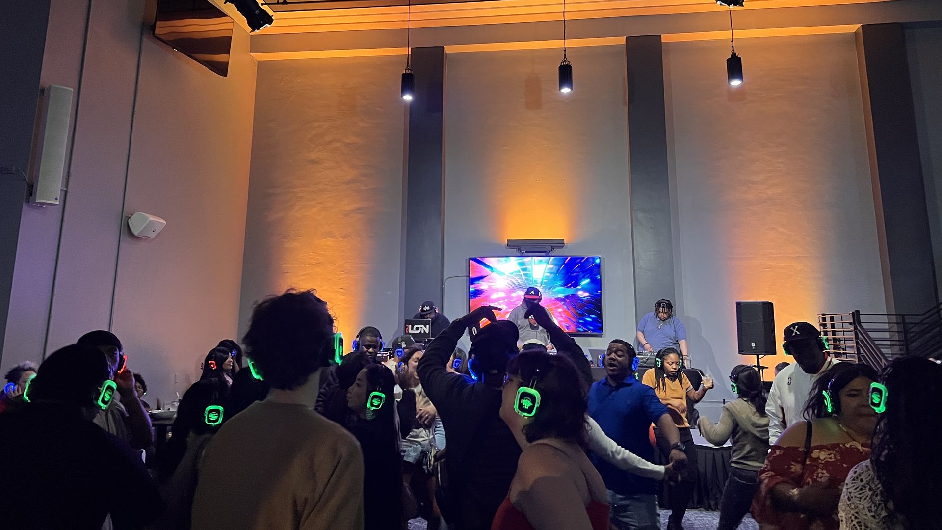 A crowd of people dancing with headphones on with three DJs in the back.