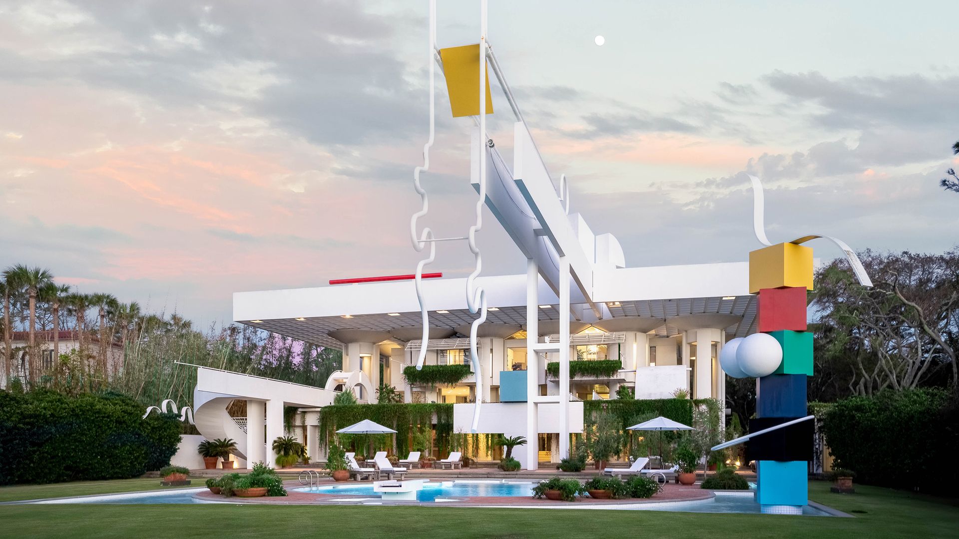 A white neo-futurist beach home with a cantilvered design element looming over a swimming pool and multicolored shape sculpture