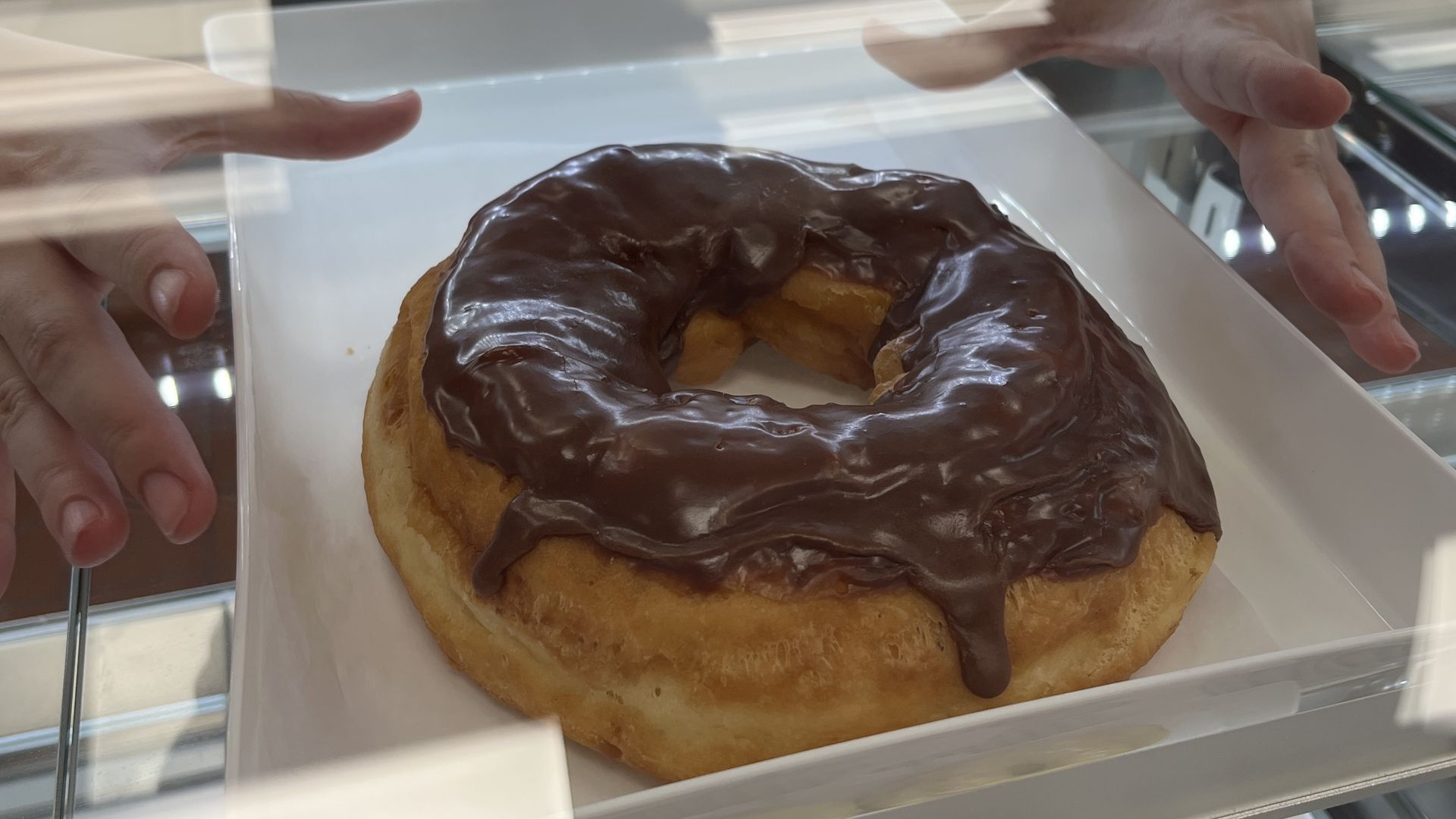 A large chocolate-glazed donut on a white tray, viewed through glass with two hands reaching towards it from either side.