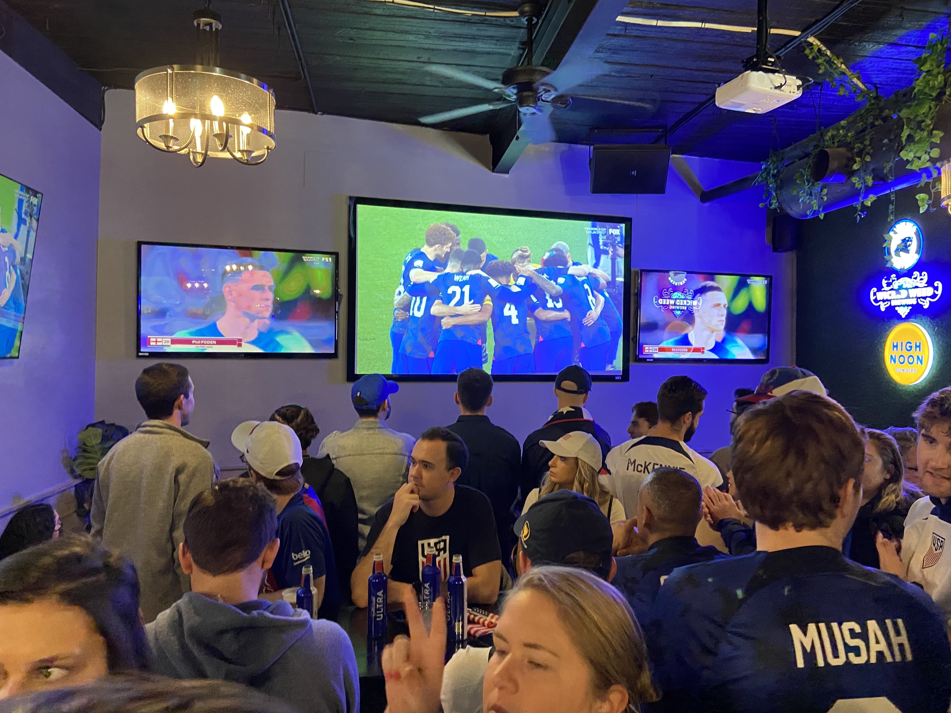 Indoor sports bar with a crowd watching a football match on three large screens; players in blue jerseys celebrate on the central screen. Blue lighting, ceiling fan, chandelier, and greenery.
