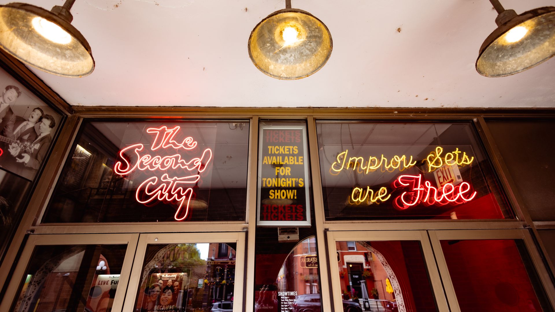 Front entrance of The Second City theater with red and yellow neon signs saying "The Second City" and "Improv Sets are Free" under three rustic ceiling lights.