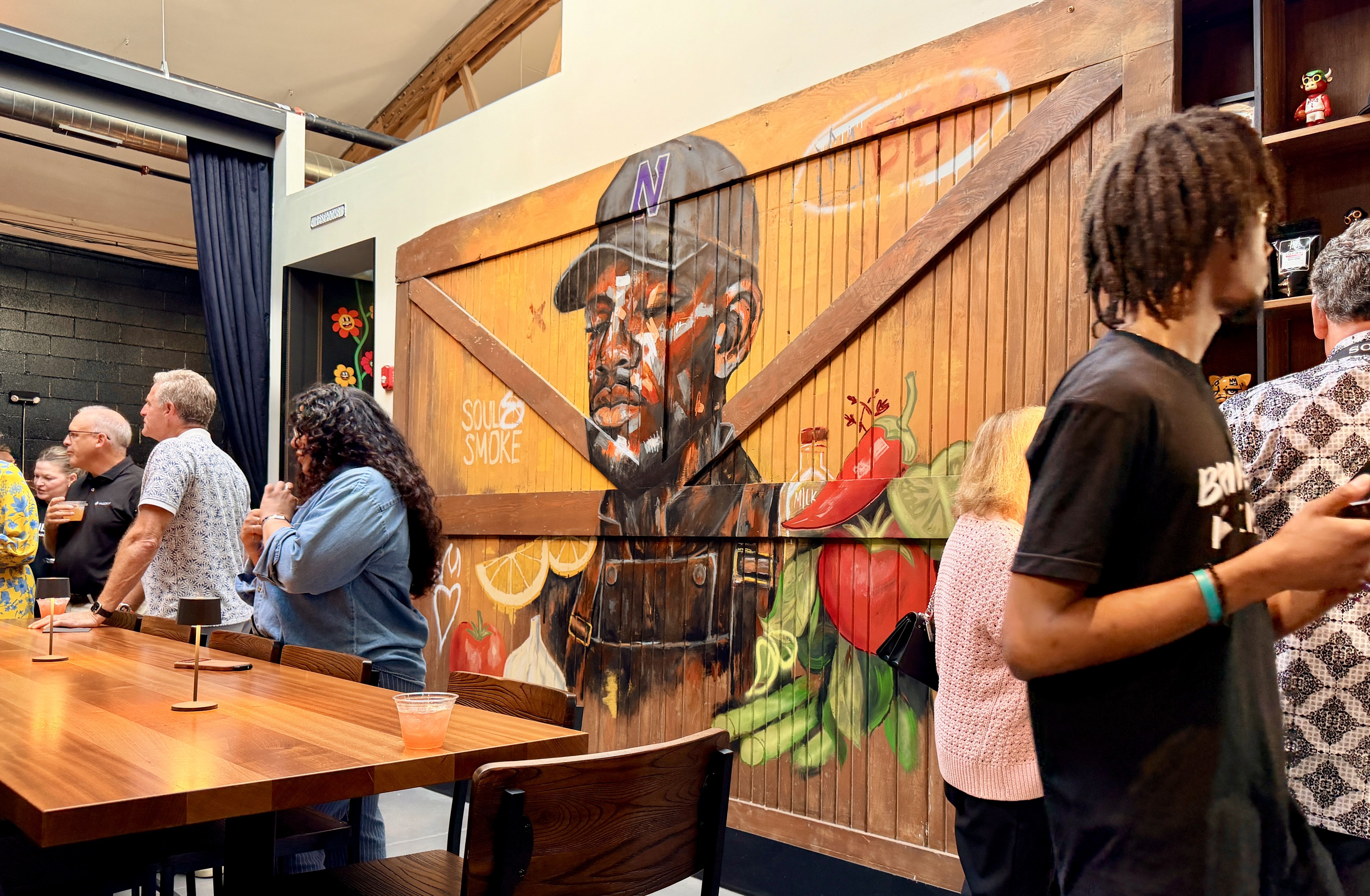 Indoor space with a large wooden mural of a man in a hat on an orange-brown wall. People stand around wooden tables with drinks; a shelf on the right holds a small figurine.