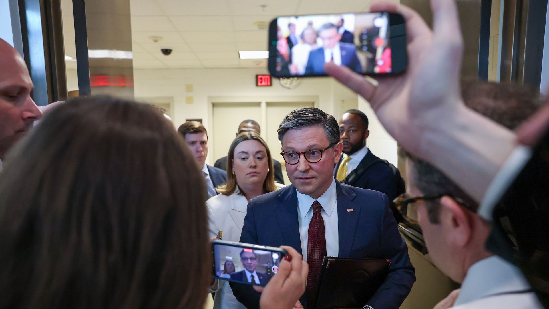 A man in a navy suit and red tie, holding a dark portfolio, walks through a hallway surrounded by reporters and photographers filming him with smartphones.