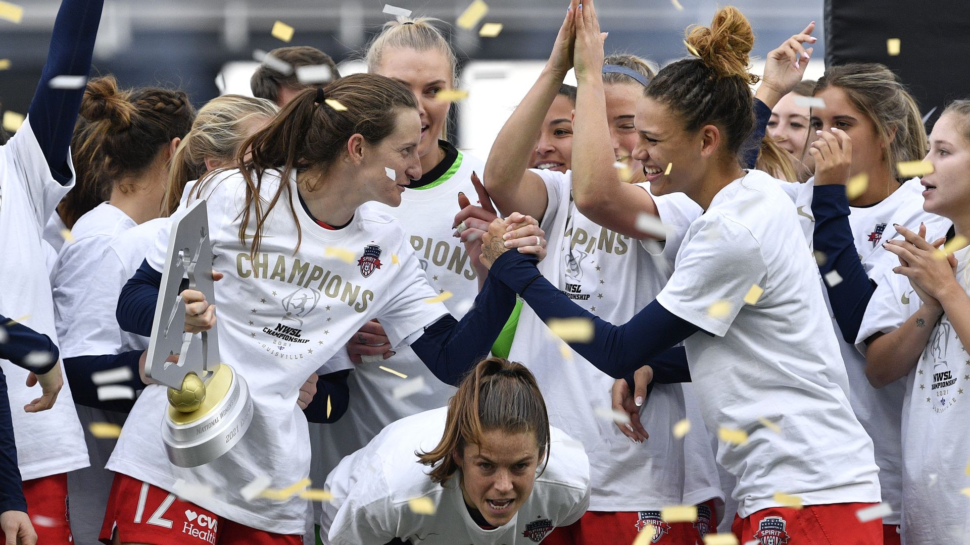 A women's soccer team celebrates, hoisting a trophy as confetti falls.