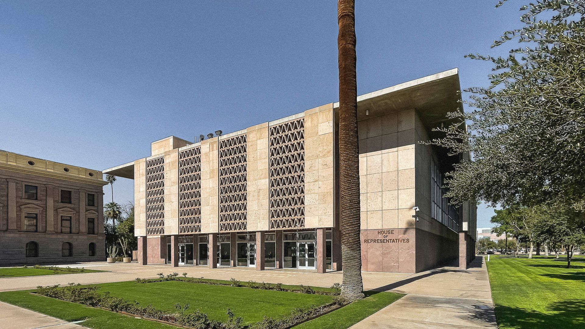 A beige building with alternating vertical solid and decorative lattice panels on the front above the doors, with a rectangle of grass and a palm tree in front; the bottom right corner reads "HOUSE OF REPRESENTATIVES".