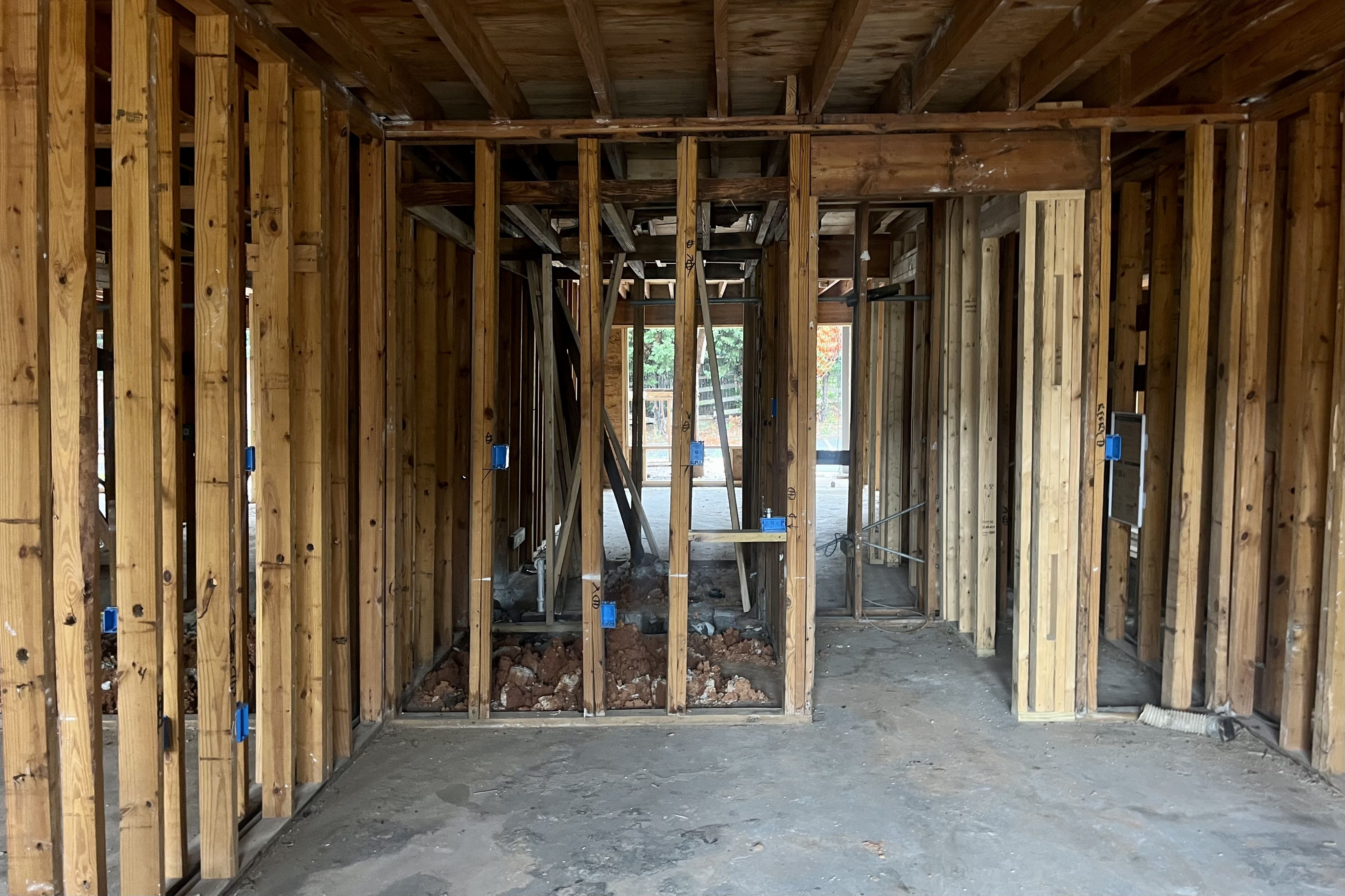 Interior view of a wooden frame under construction with exposed studs, electrical boxes, and trenches dug in the concrete floor.