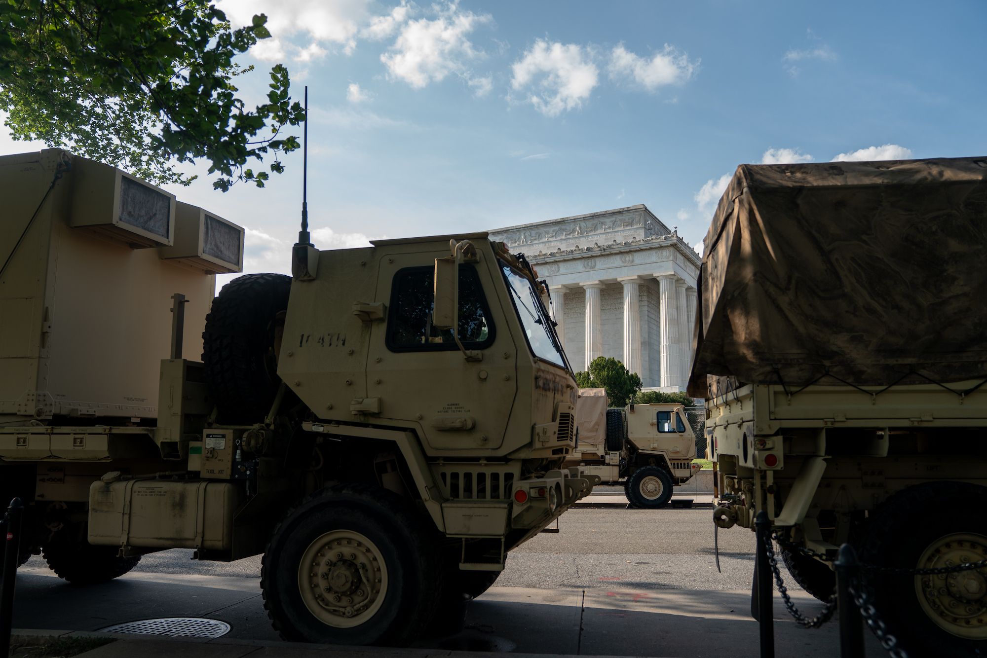 Tan military trucks parked on a street with the white Lincoln Memorial and a blue sky with clouds in the background.