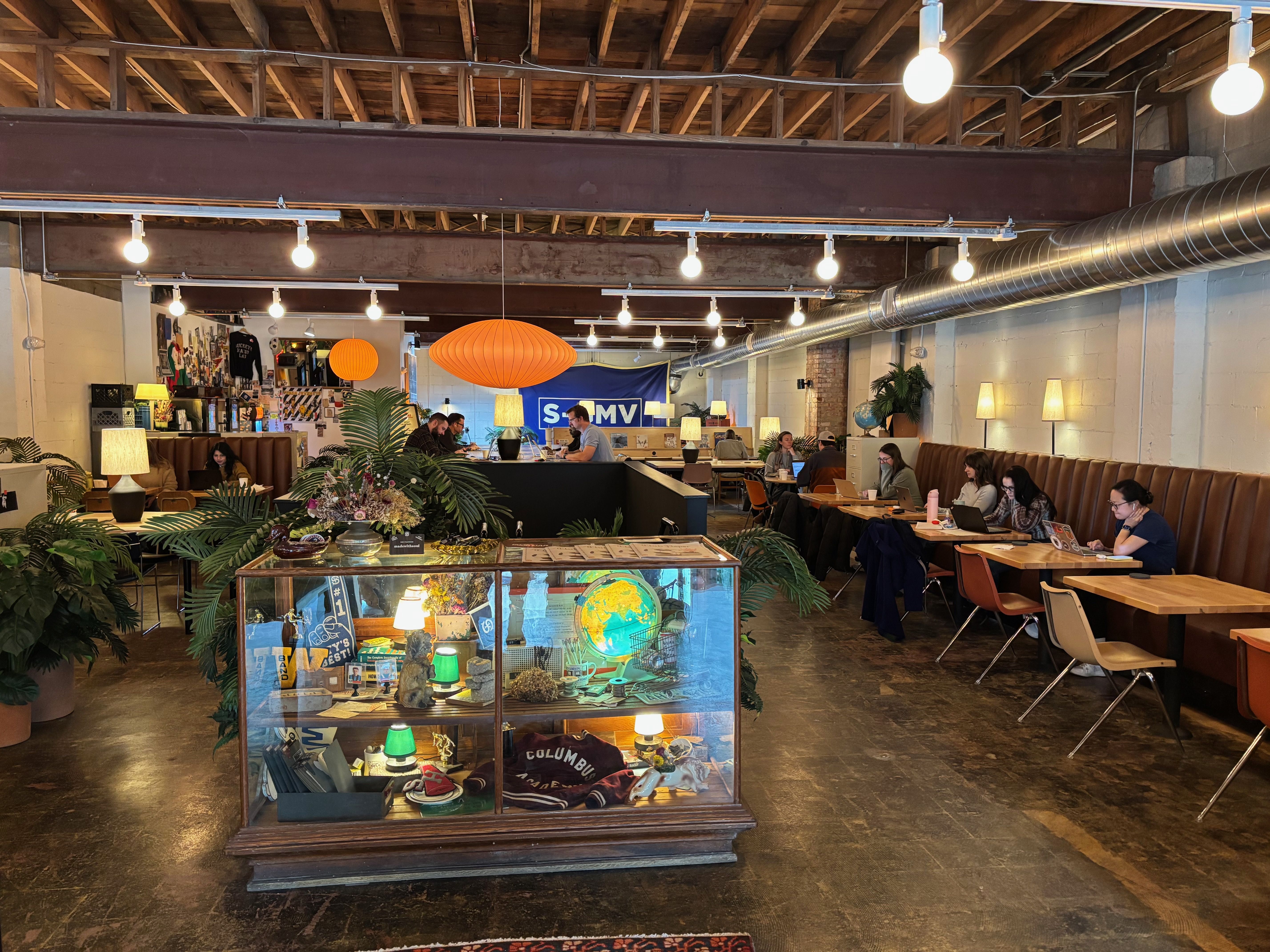 Industrial coworking cafe with exposed ceiling beams, string lights, and orange lanterns. A glass display cabinet sits in the foreground while people work at wooden tables along a long bench.