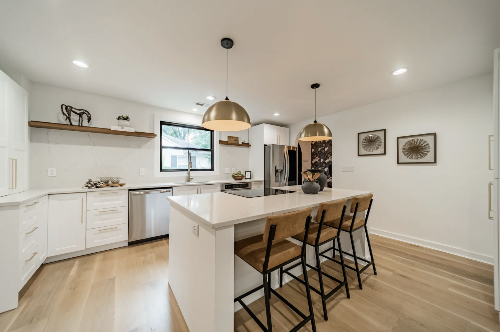 Bright modern kitchen with white cabinets, marble backsplash, and a large island. Three brown stools line the island under brass pendant lights; stainless appliances, light wood floors, and wall art.
