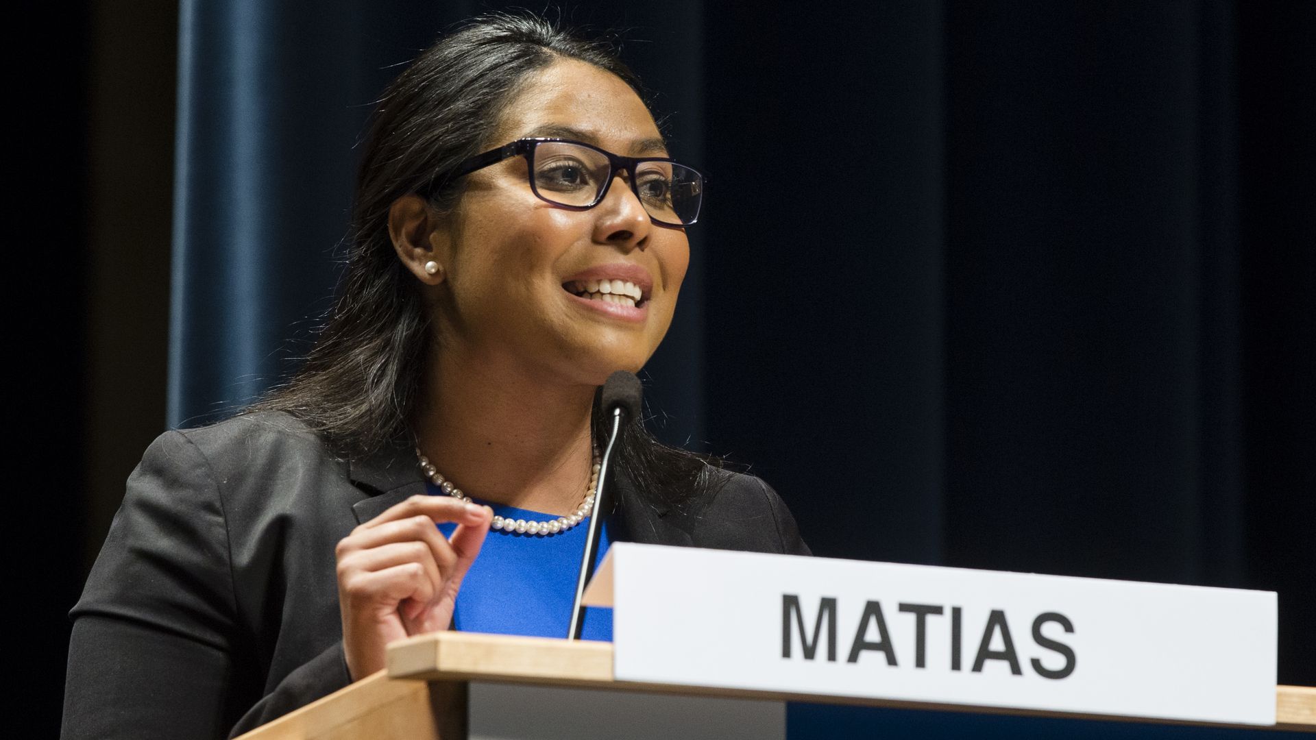 Woman with glasses, pearl earrings, and necklace speaking at a podium labeled "MATIAS," wearing a black blazer and blue top, with a dark blue curtain background.
