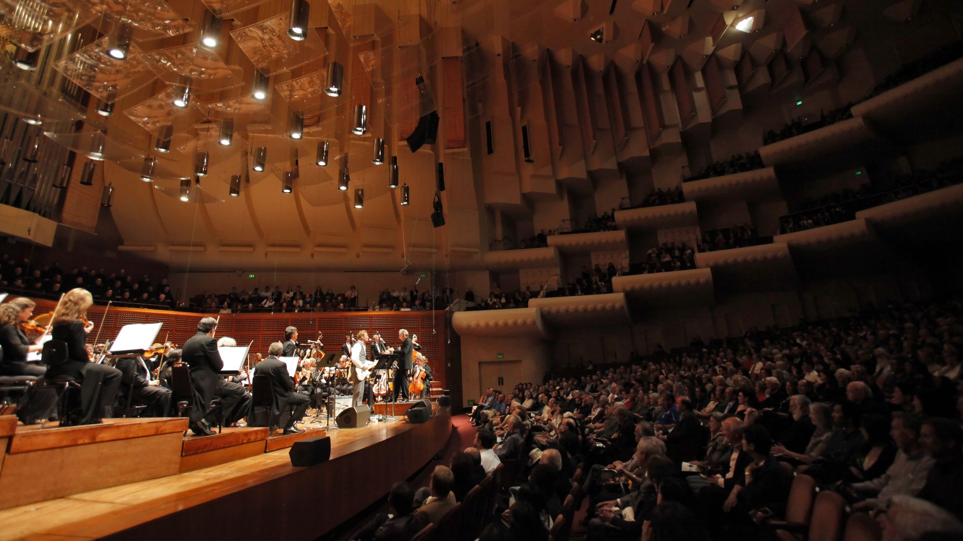 A large concert hall with an orchestra on stage, musicians playing strings under warm lighting as a full audience fills the tiered seats in the dim auditorium.
