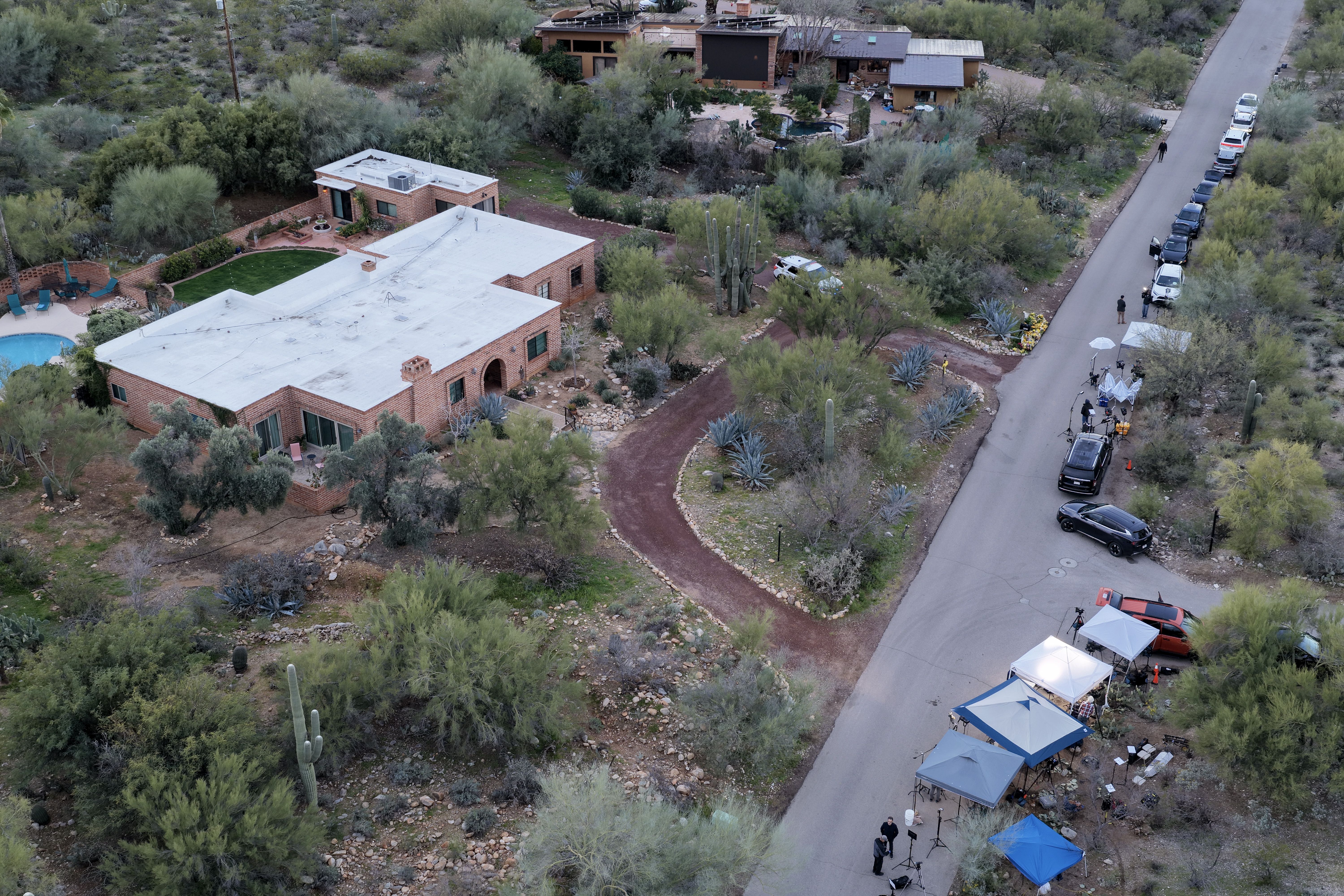 Aerial view of a desert neighborhood with brick houses and vegetation, a line of parked cars, and several blue and white tents set up beside the road, suggesting a filming or event setup.