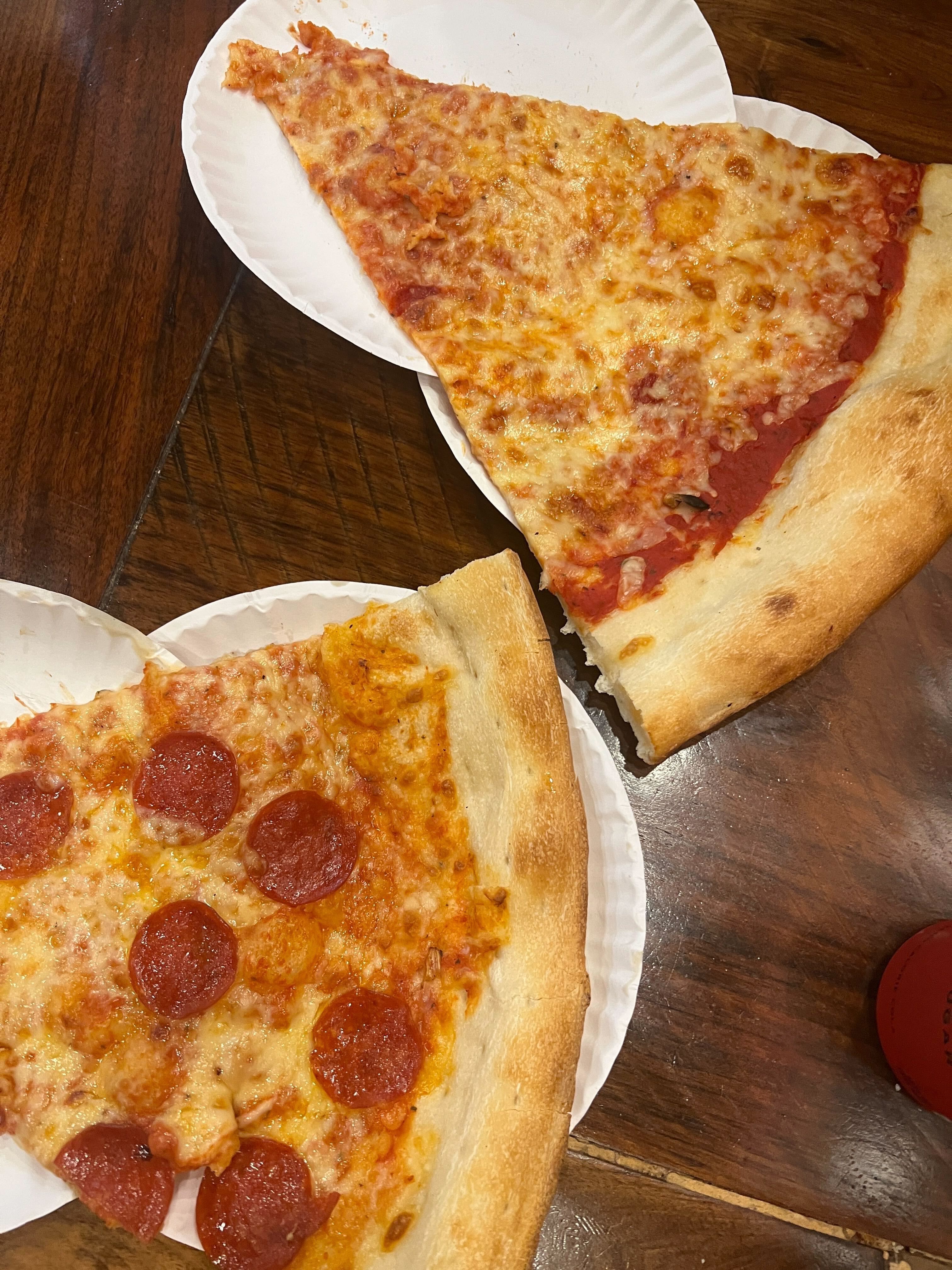 Two large pizza slices on white paper plates on a wooden table; one slice is plain cheese, the other has pepperoni.