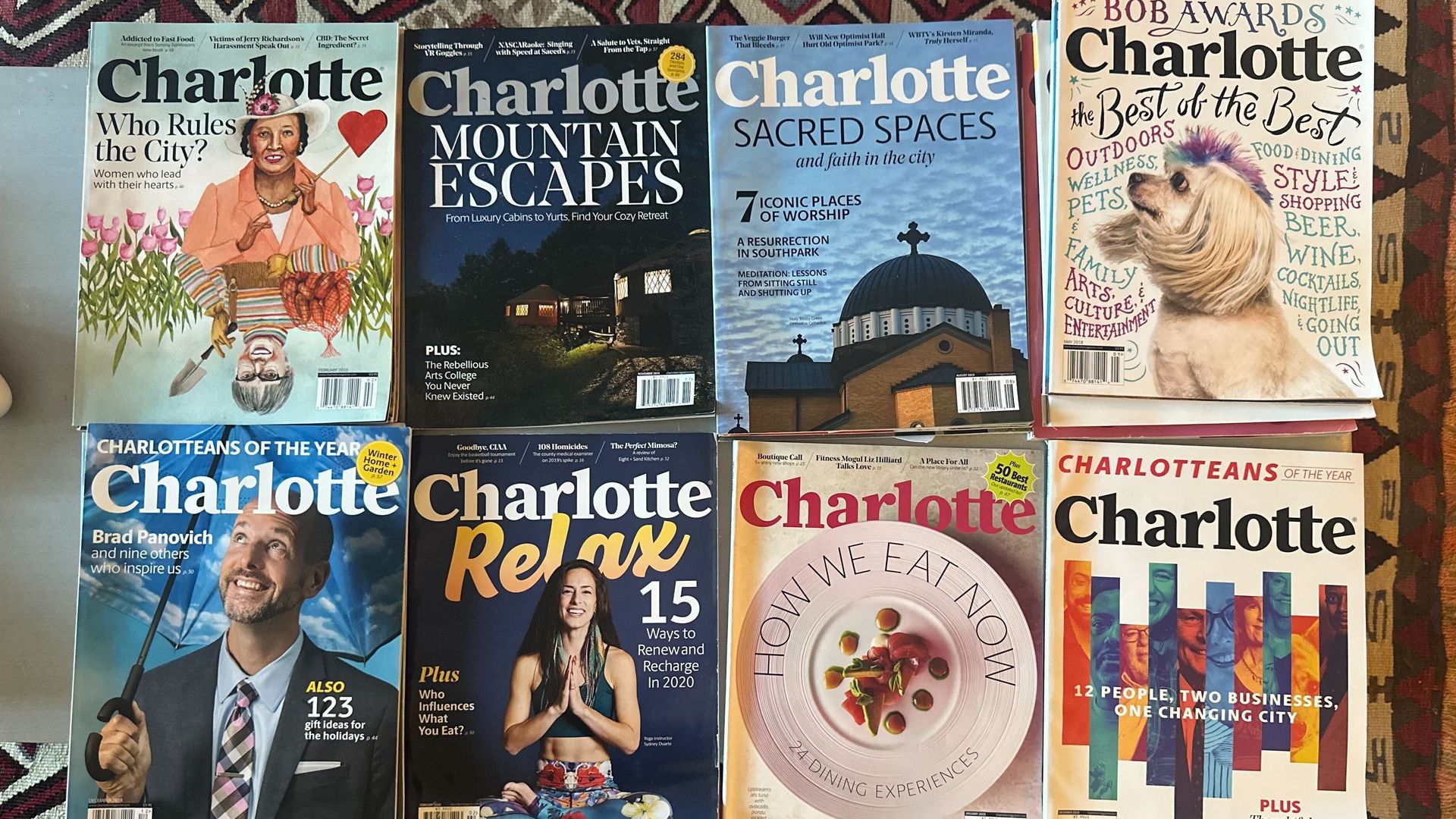 Eight Charlotte magazine covers arranged on a patterned rug, featuring topics like city leadership, mountain escapes, sacred spaces, awards, people profiles, relaxation, dining, and holiday gifts.
