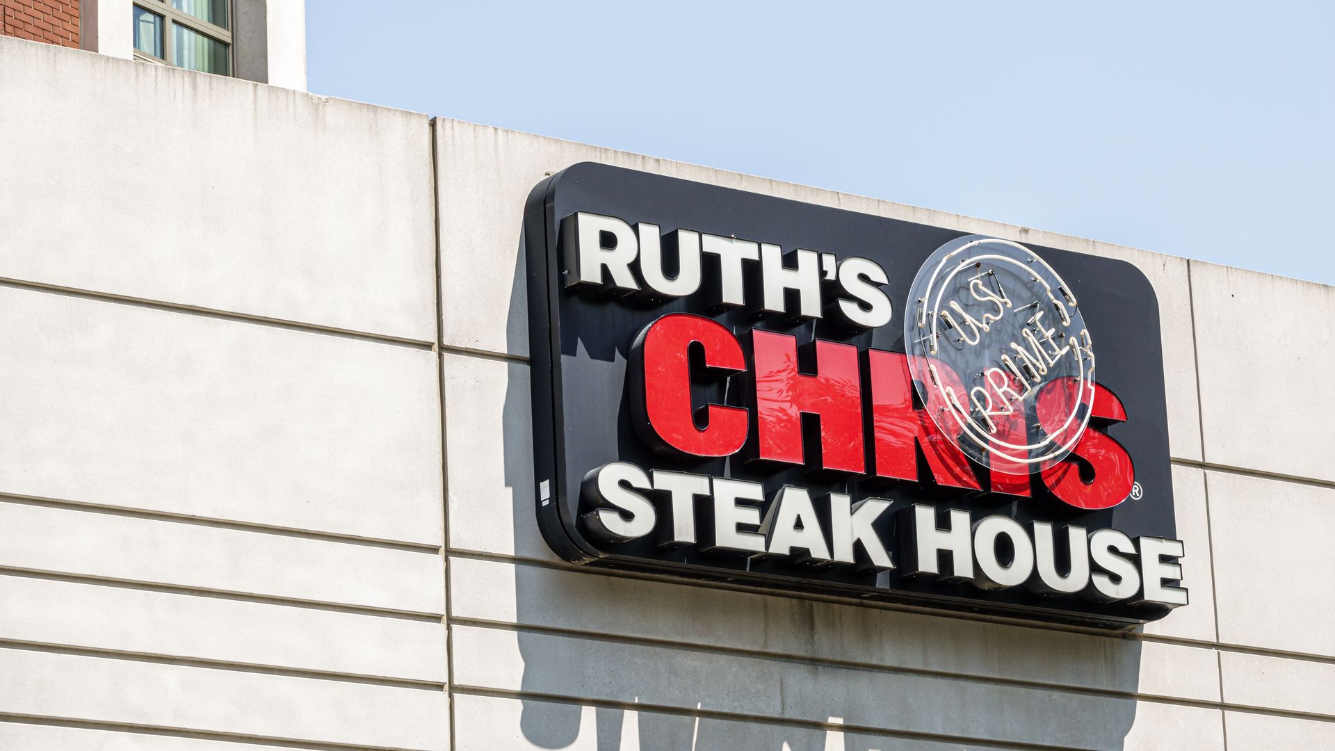 Exterior view of Ruth's Chris Steak House sign mounted on a light gray building, with bold white letters and bright red CHRIS, plus a circular neon emblem, against a blue sky.