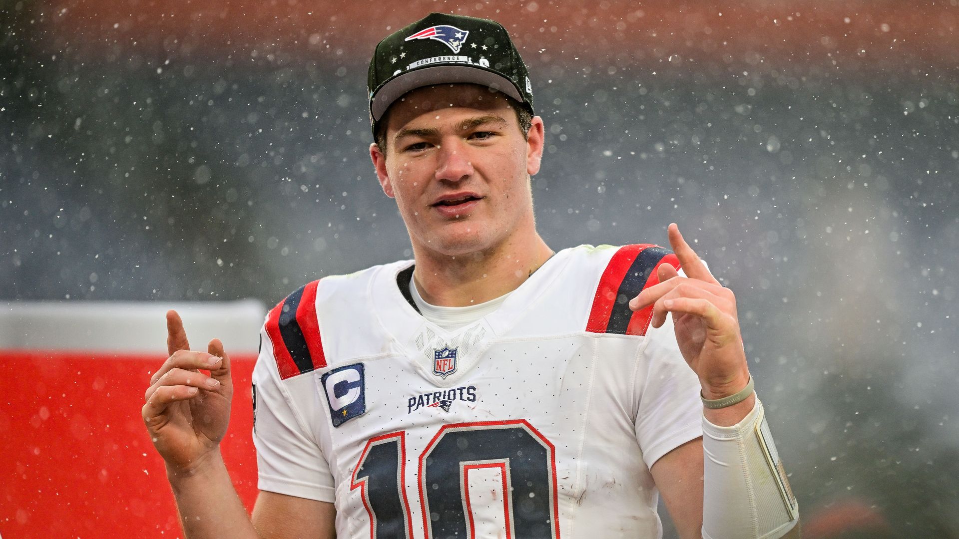 New England Patriots quarterback Drake Maye looks on after a win against the Denver Broncos in the AFC Championship Game. It is snowing. He is pointing up with both hands.