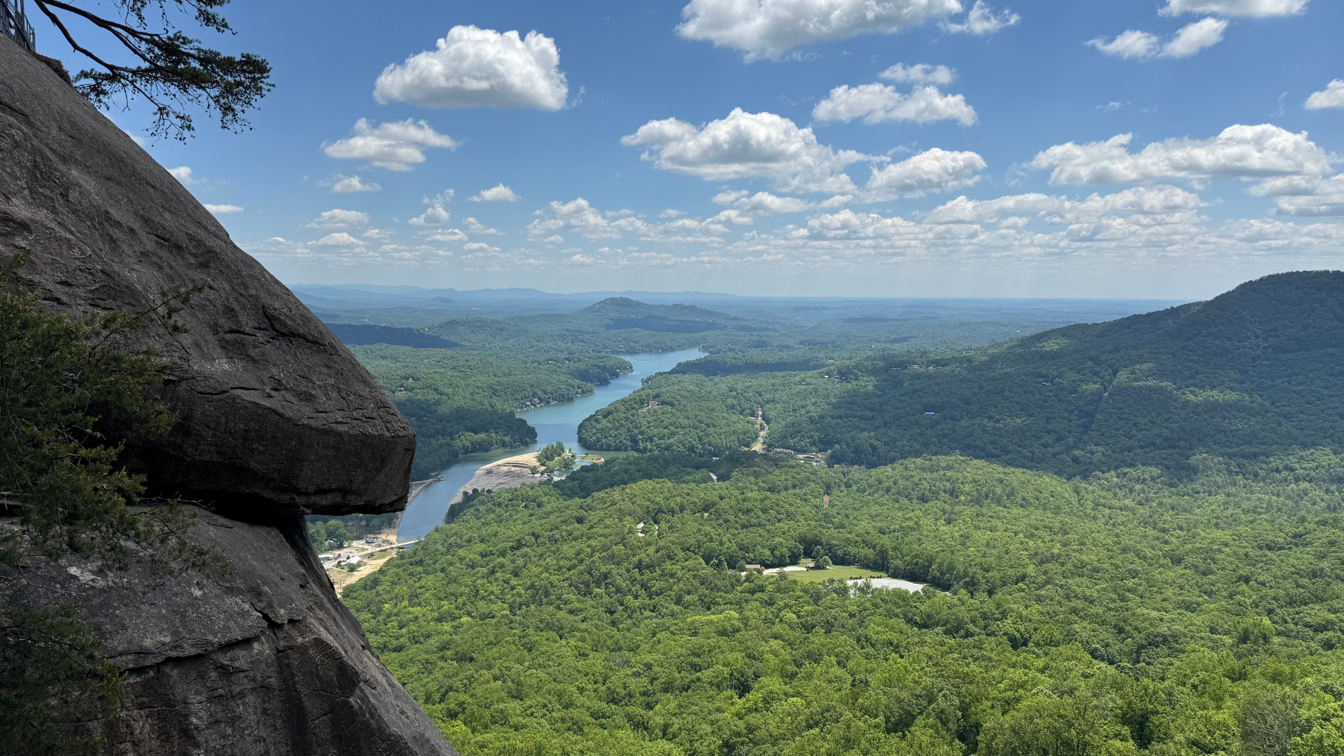 View from a rocky cliff on the left shows a wide green valley, a winding river, distant hills, and a bright blue sky with scattered white clouds.