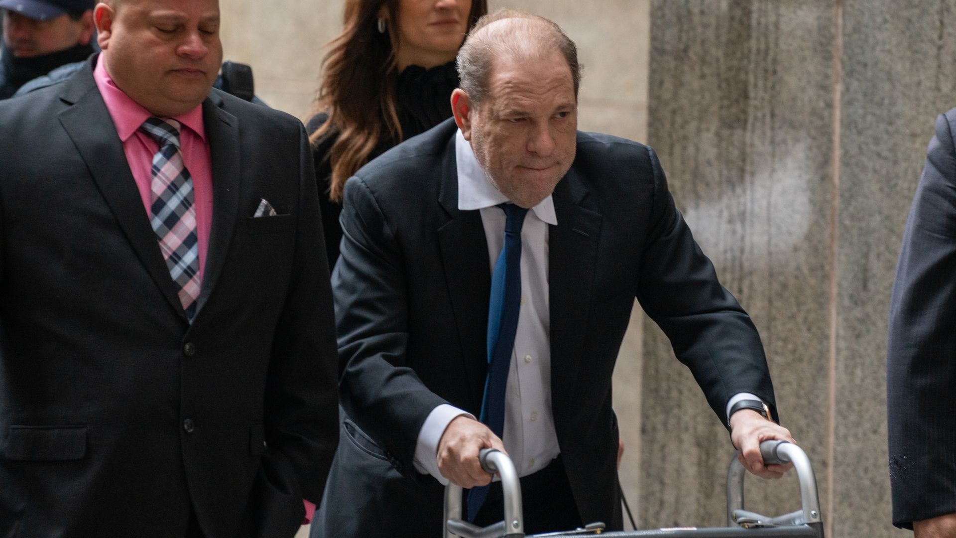 In this image, Harvey Weinstein uses a walker to walk down a hallway while wearing a suit and tie