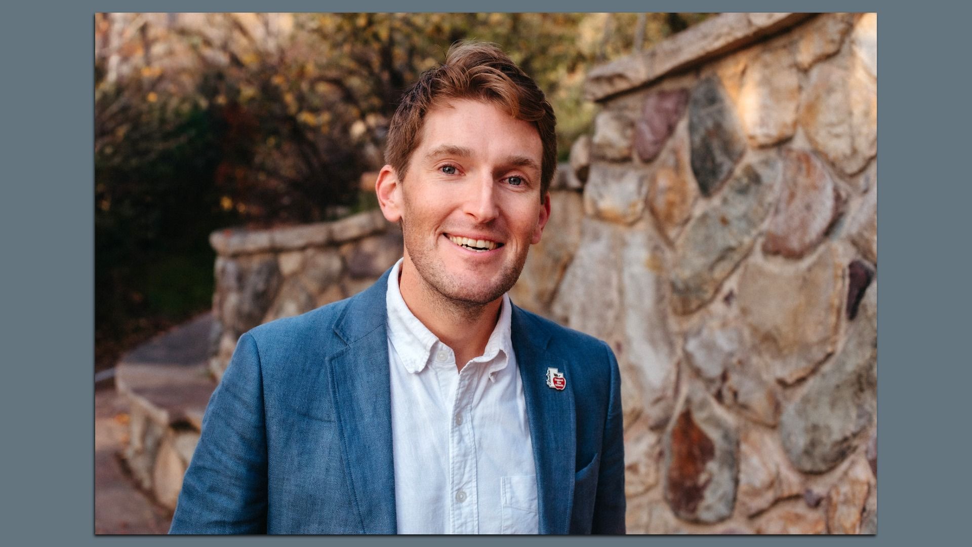 Smiling young man with light brown hair wearing a blue blazer and white shirt stands outdoors near a stone wall with autumn foliage in the background.