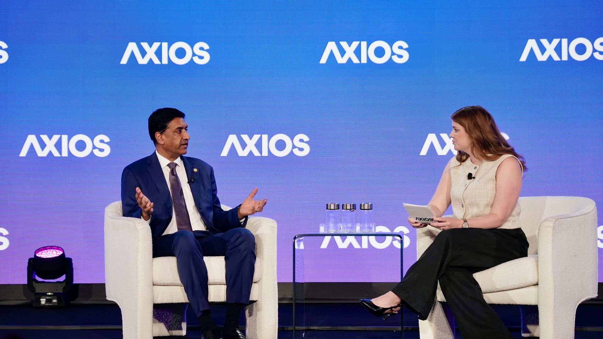 Two people seated on white chairs on a stage with a blue backdrop displaying "AXIOS" logos. The man in a navy suit speaks while the woman in a beige sleeveless top and black pants listens, holding notes.