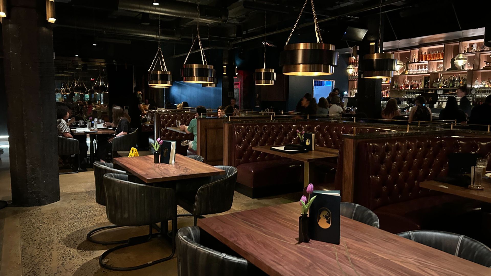Dimly lit restaurant interior with dark leather booths, wooden tables, hanging black-and-gold lamps, and patrons seated and conversing near a bar stocked with bottles.