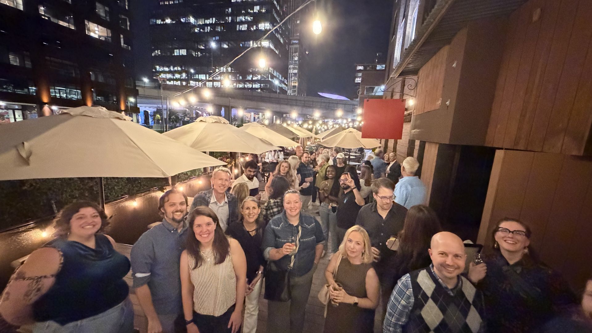 Nighttime rooftop terrace party with string lights and beige umbrellas; a large crowd smiles and poses for the camera, with tall city buildings lit up in the background.