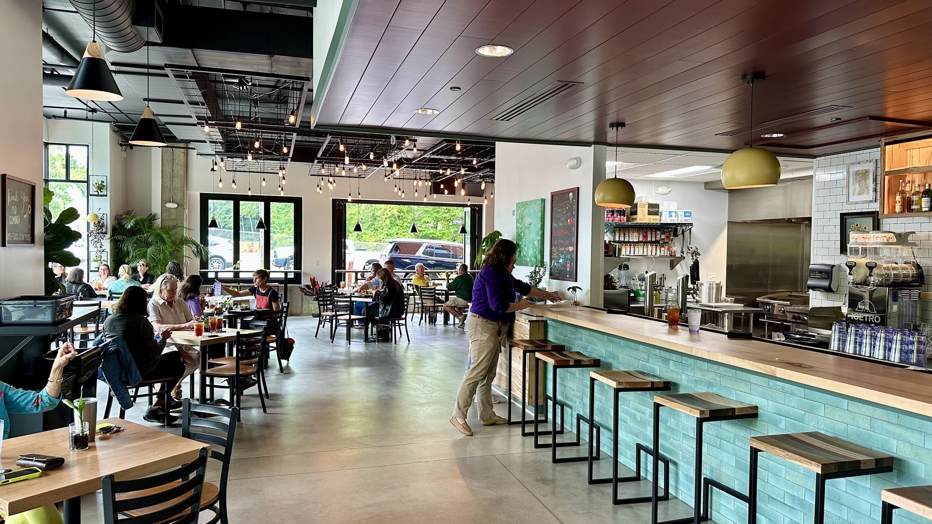 Inside a modern cafe, teal-tiled counter with wooden stools runs along the right. Customers sit at tables beneath a high, exposed-ceiling with ductwork and hanging bulbs; large windows show greenery outside.