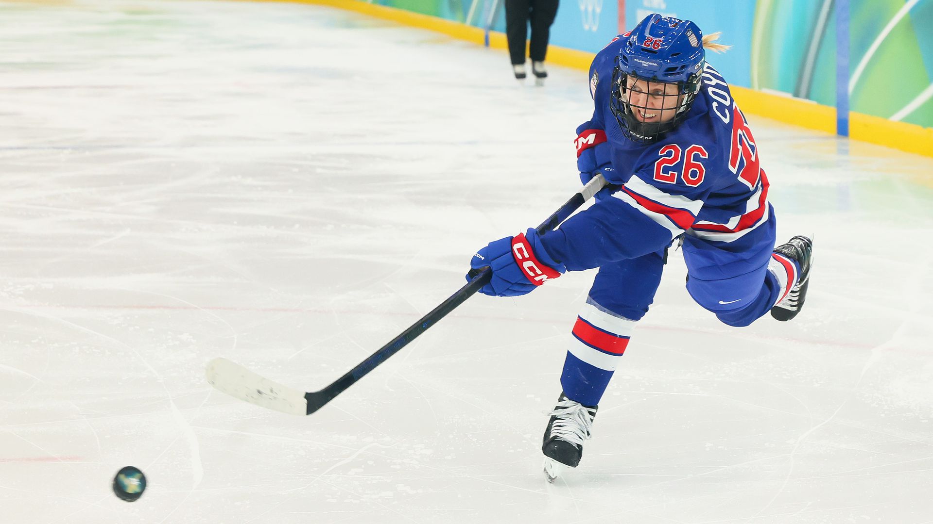 Ice hockey player in blue uniform with number 26 shooting puck on ice rink, focused expression, red and white stripes on jersey and gloves, colorful rink boards in background.