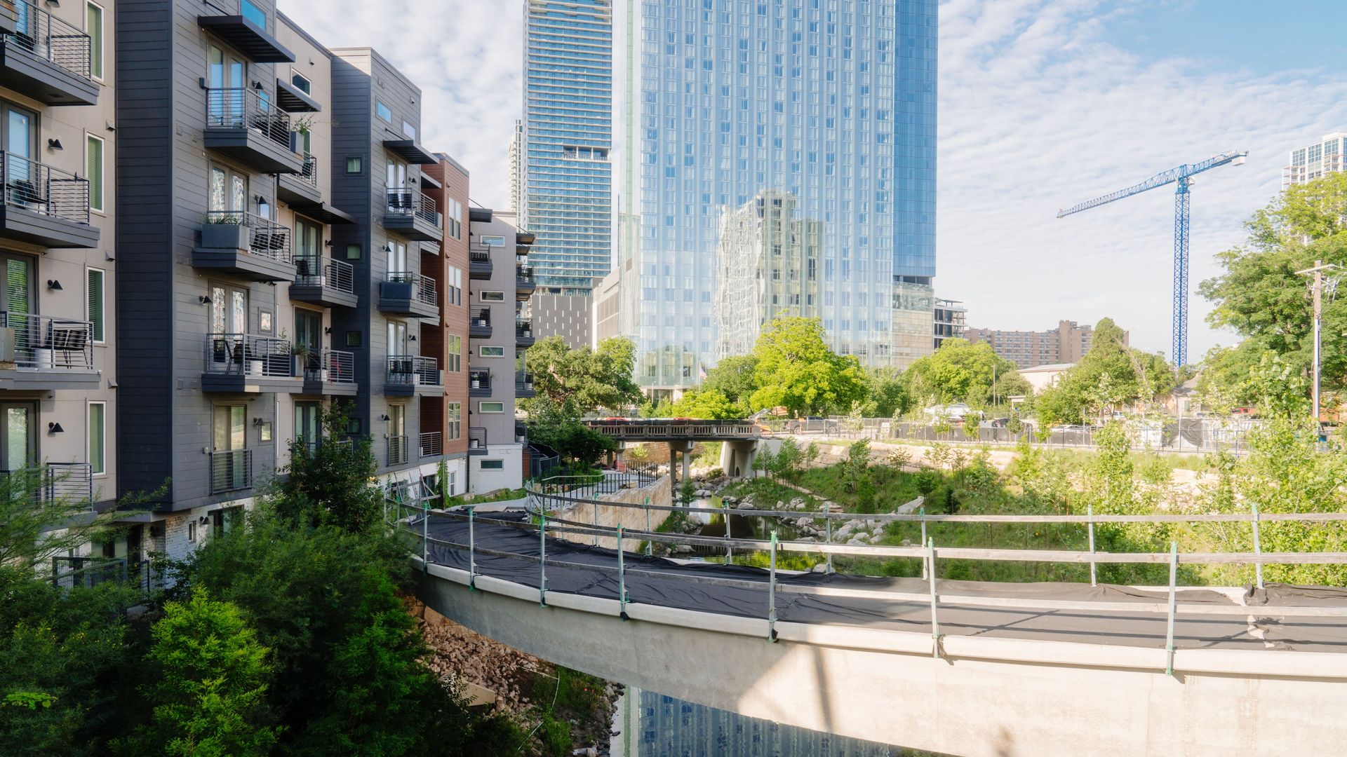 Row of modern apartments with balconies on the left, a tall glass skyscraper center, a blue crane on the right, and a walkway over a reflective canal with trees and sky.