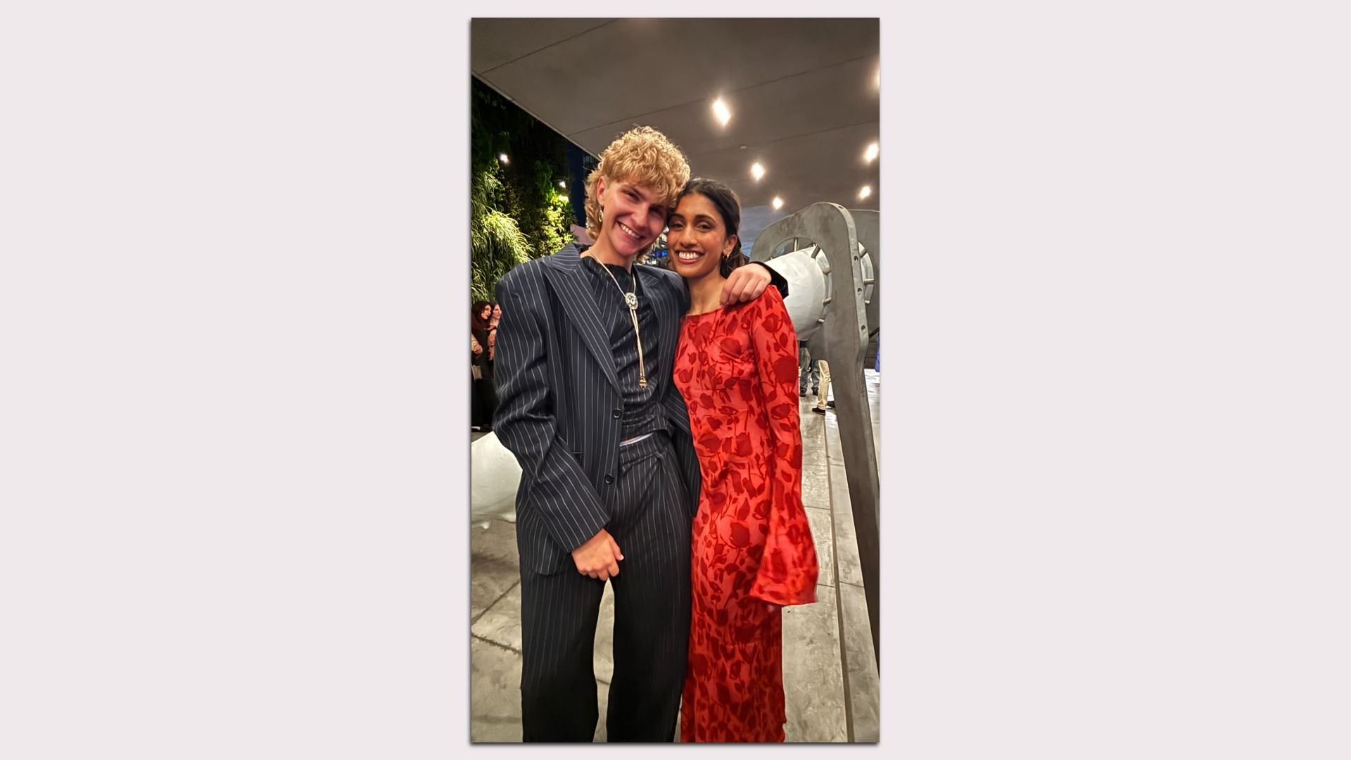 Two friends pose indoors: a blond person in a pinstripe suit with a necklace and a woman in a red floral dress, smiling with arms around each other, plants and a metallic sculpture in the background.