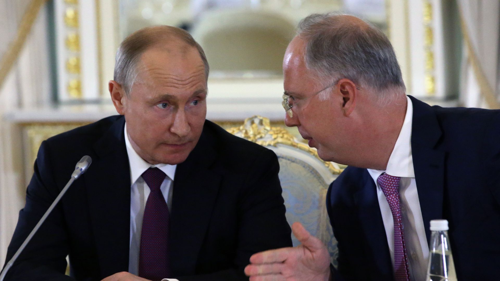 A balding, gray-haired fair-skinned Russian leader Vladimir Putin, wearing a black suit jacket, white shirt and mauve tie, and a balding, gray-haired fair-skinned and bespectacled Russian businessman Kirill Dmitriev, wearing a black suit jacket, white shirt and purple tie, speaking at a table 