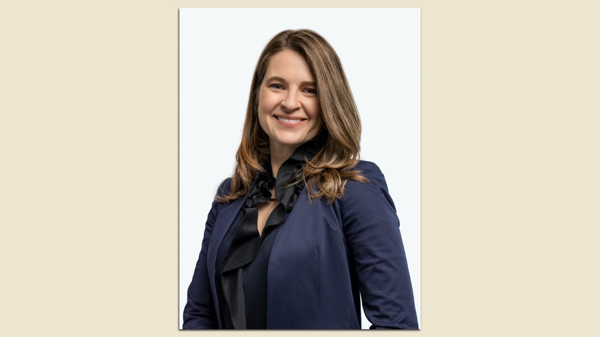 Portrait of a smiling woman with shoulder-length brown hair, wearing a navy blazer and black blouse, posed at a slight angle against a light gray backdrop.