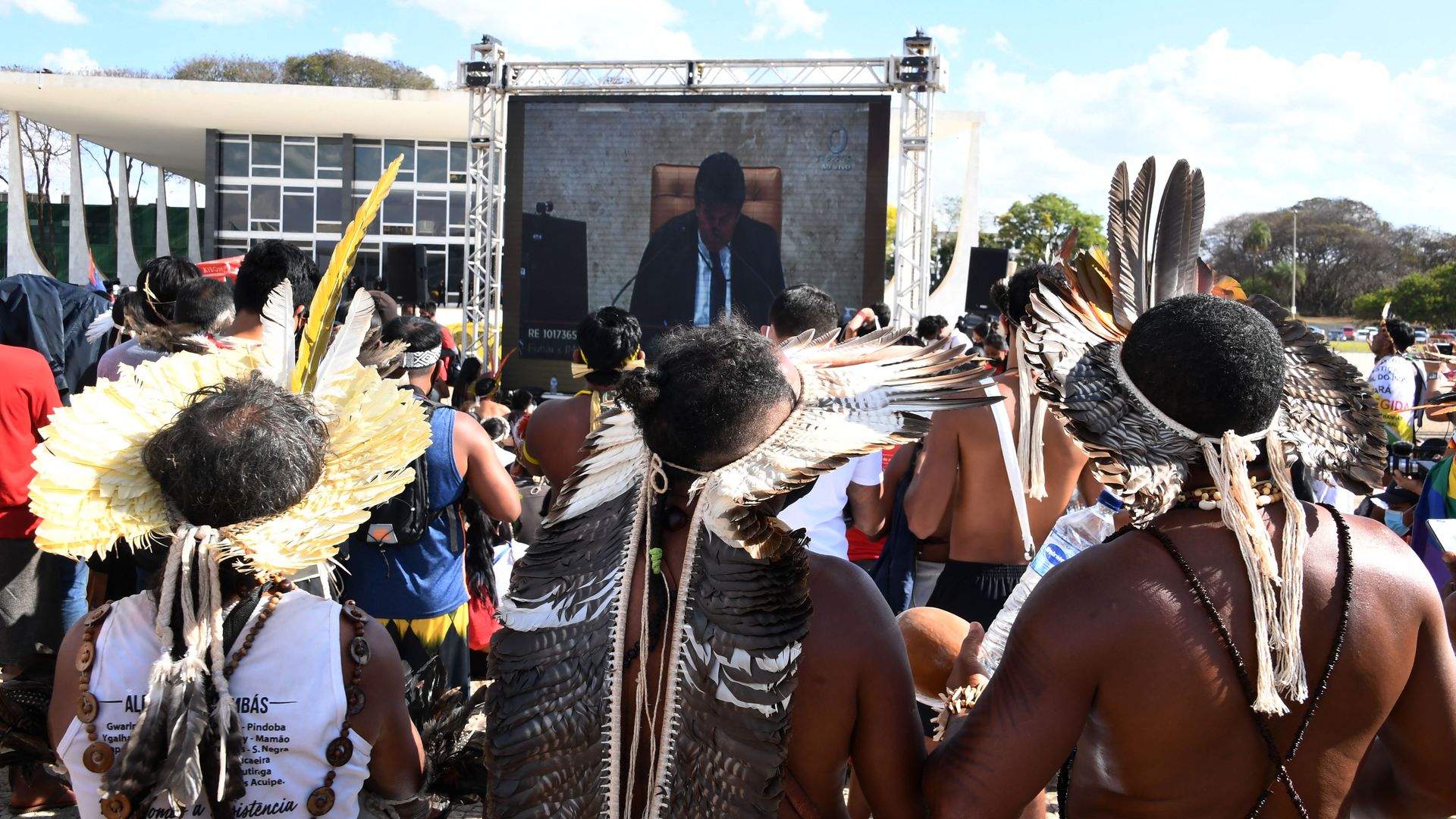 Two Indigenous Brazilian men watch a large outdoor screen with President Jairo Bolsonaro. 