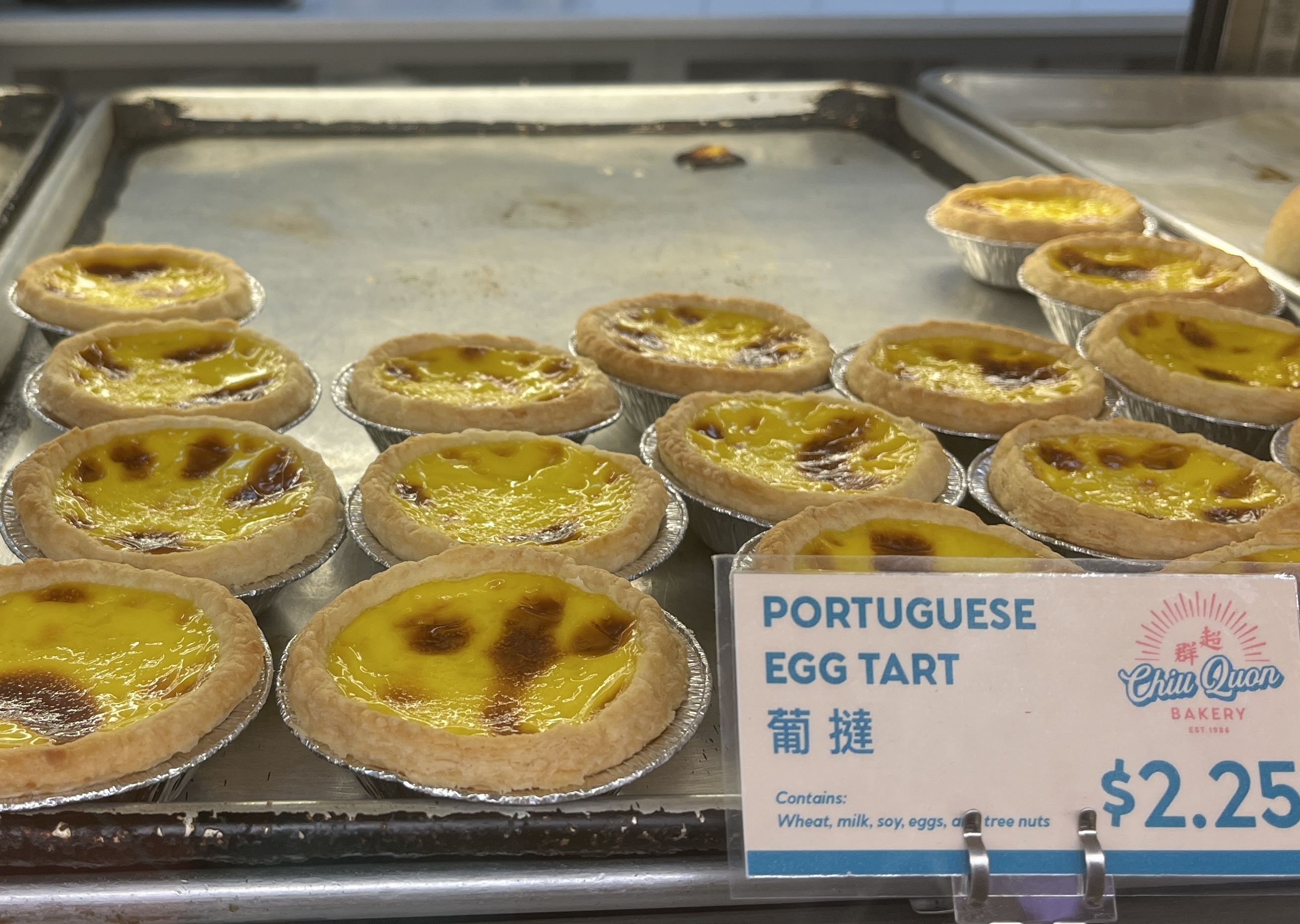 Tray of Portuguese egg tarts with browned tops in foil cups at Chiu Quon Bakery, priced $2.25 each. Sign notes ingredients include wheat, milk, soy, eggs, and tree nuts.