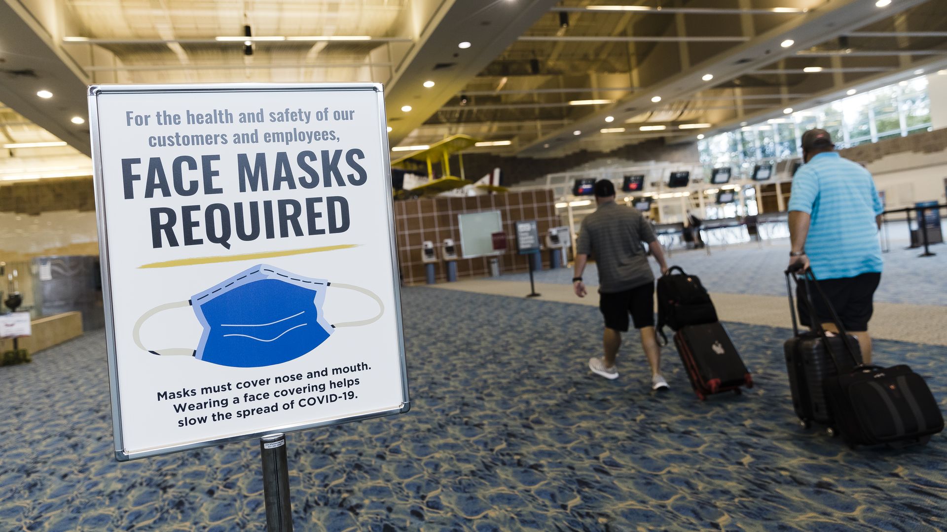 Photo of a sign that says "Face masks required" in an airport with two travelers on the right
