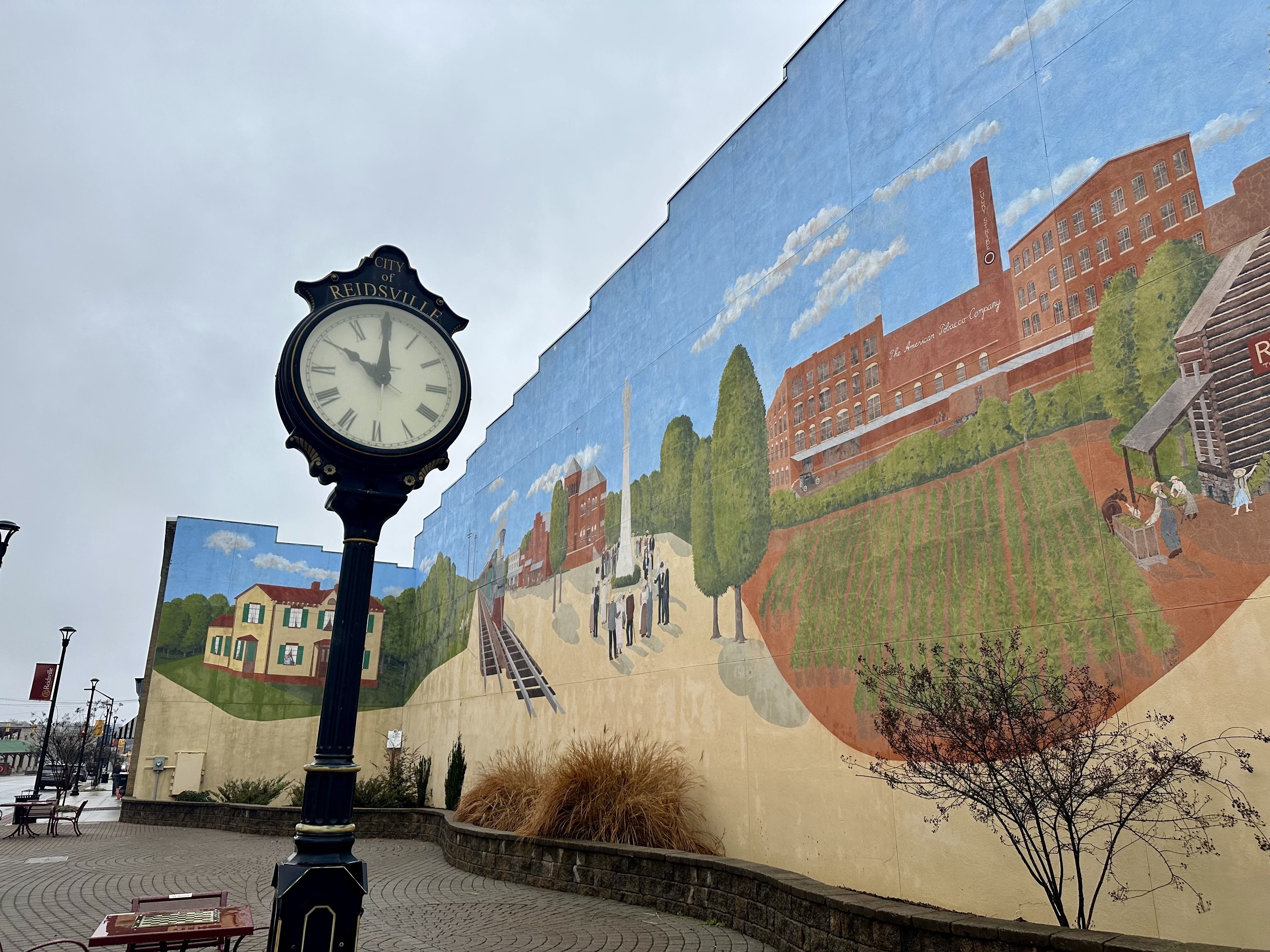 Black street clock showing 11:55 in front of a large mural depicting historic scenes including buildings, trees, people, and railroad tracks under a cloudy sky in Reidsville.