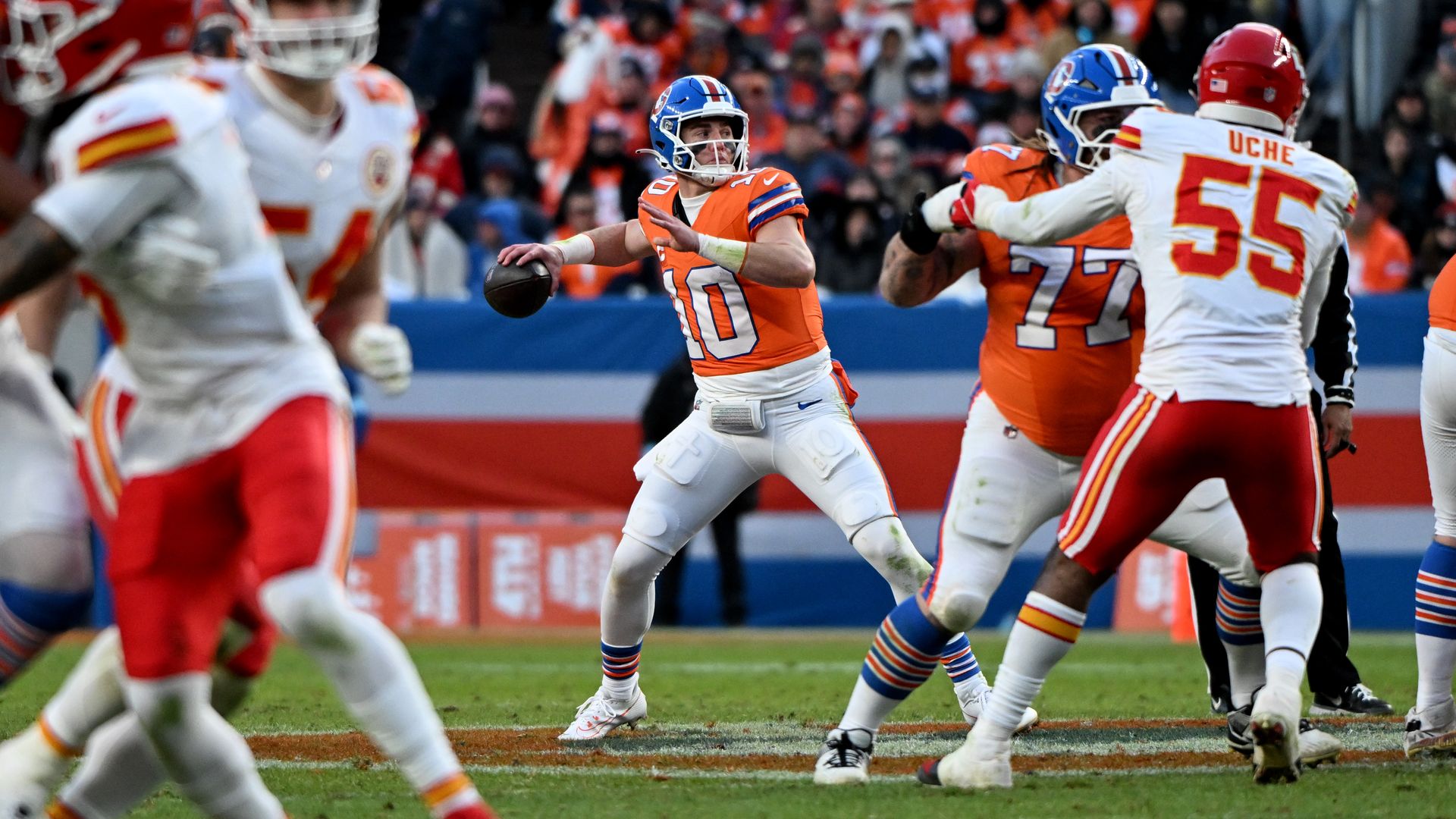 A man in a blue helmet and orange shirt prepares to throw a football while surrounded by other men in similar uniforms. 