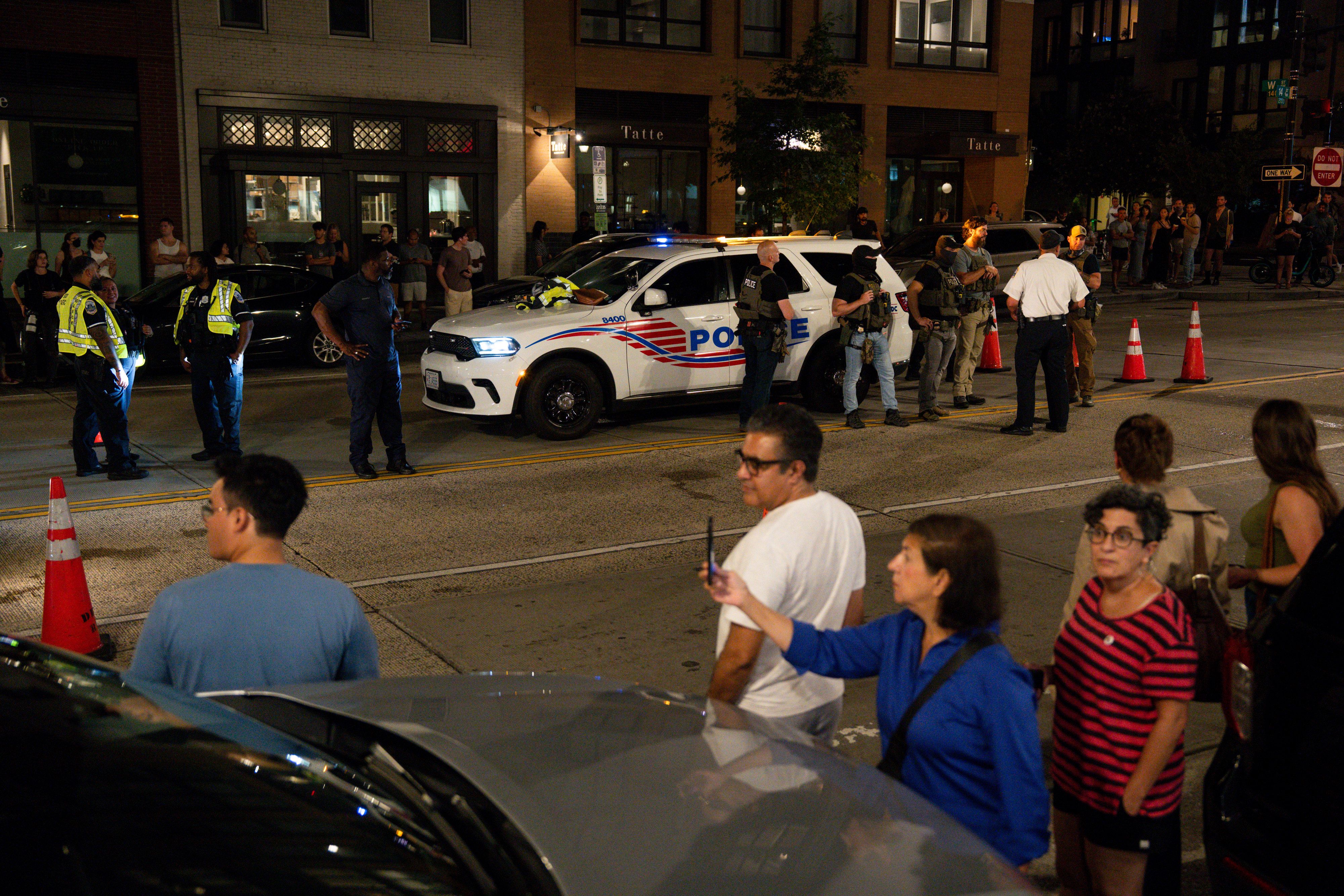 Night scene of a street with police officers in uniforms and tactical gear standing near a police SUV with flashing lights, nearby crowd of onlookers and orange traffic cones.