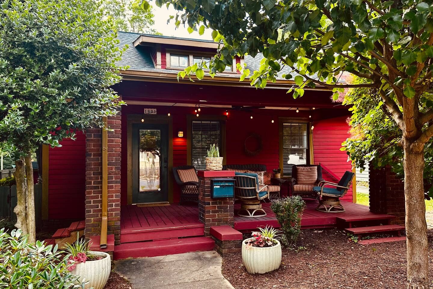 Red wooden house with a covered front porch featuring outdoor chairs and string lights, flanked by trees and potted plants in cream-colored pots on a mulched yard.