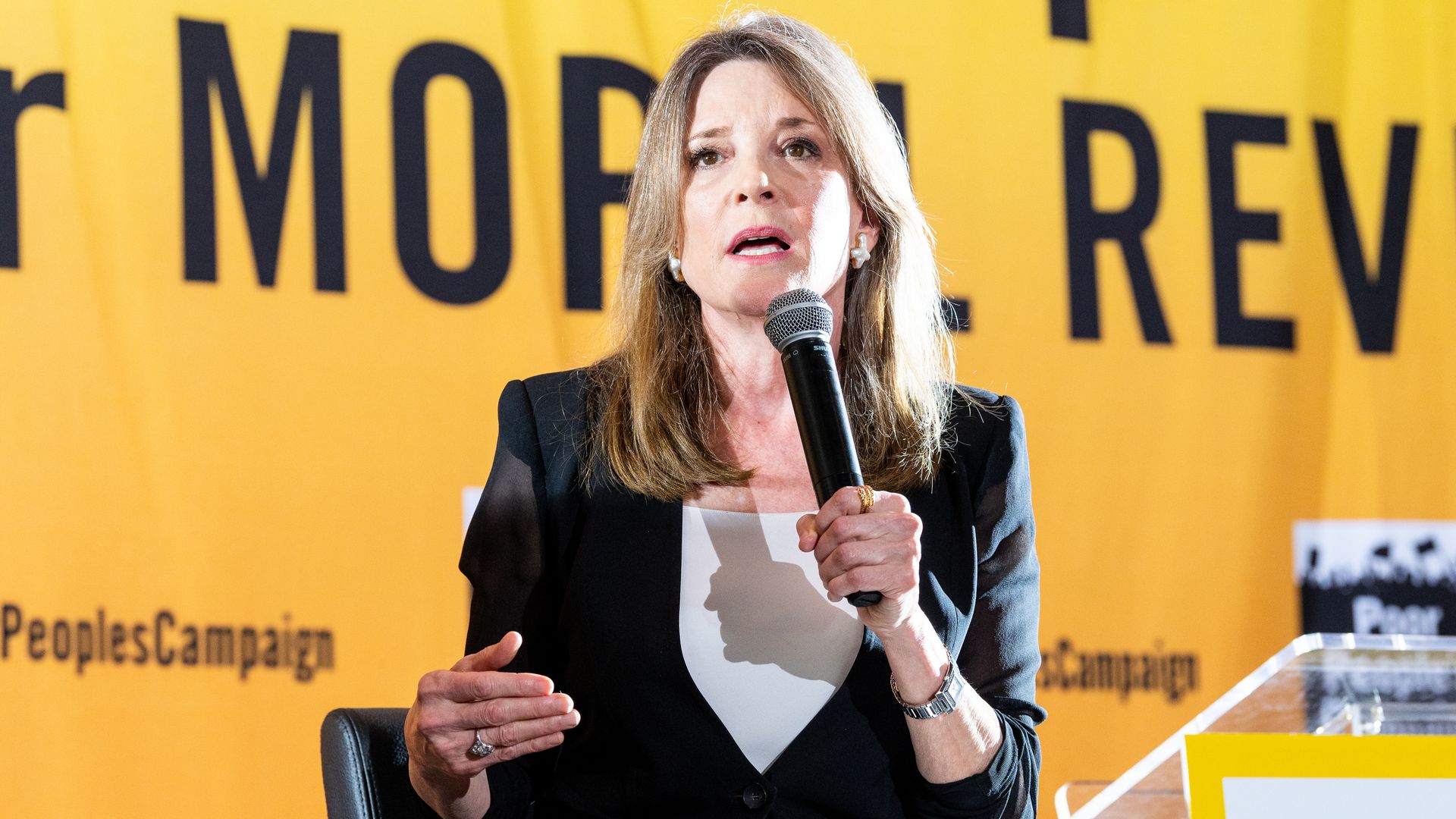 Marianna Williamson speaking into a microphone. She is wearing a black blazer with a white shirt and sitting in a leather-looking chair. 