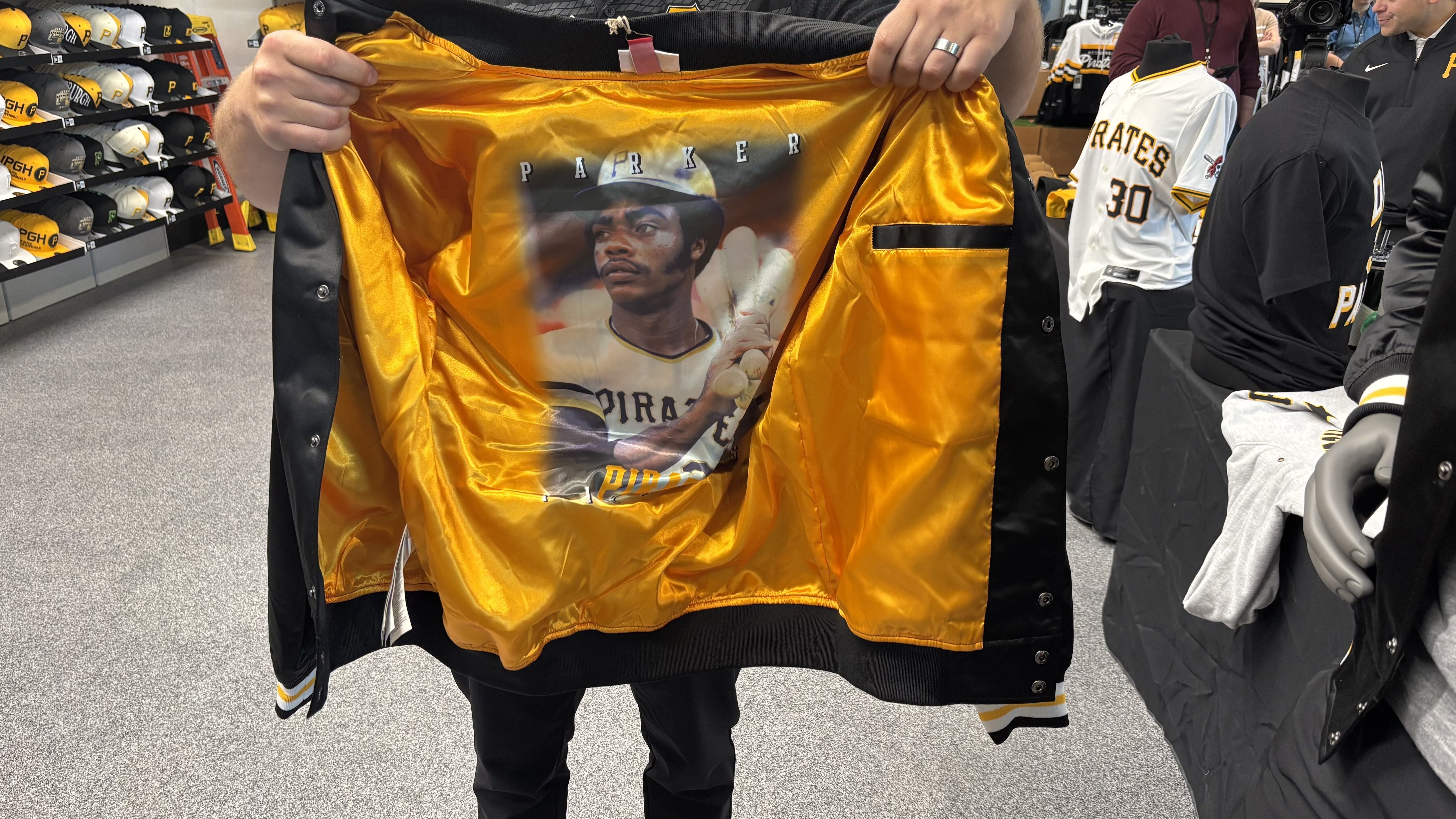 Person in a store holds open a shiny gold satin jacket to reveal a large inner image of a baseball player in a Pirates uniform with the word Parker above; Pirates hats line shelves in the background.