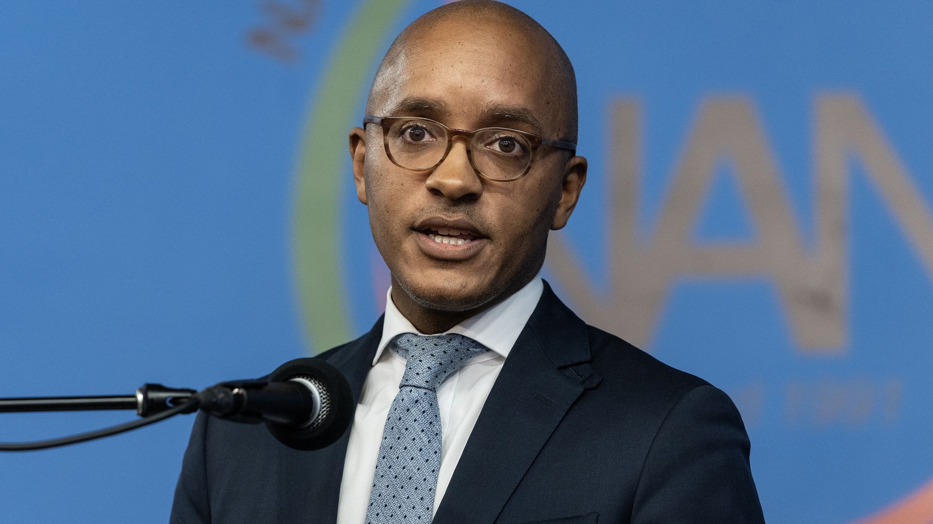 Man in suit with blue tie speaks at press conference.