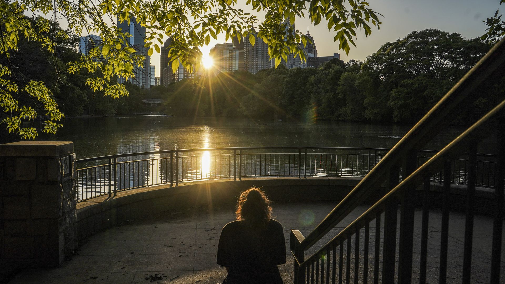 An individual sits on stairs facing a lake at a park in an urban area