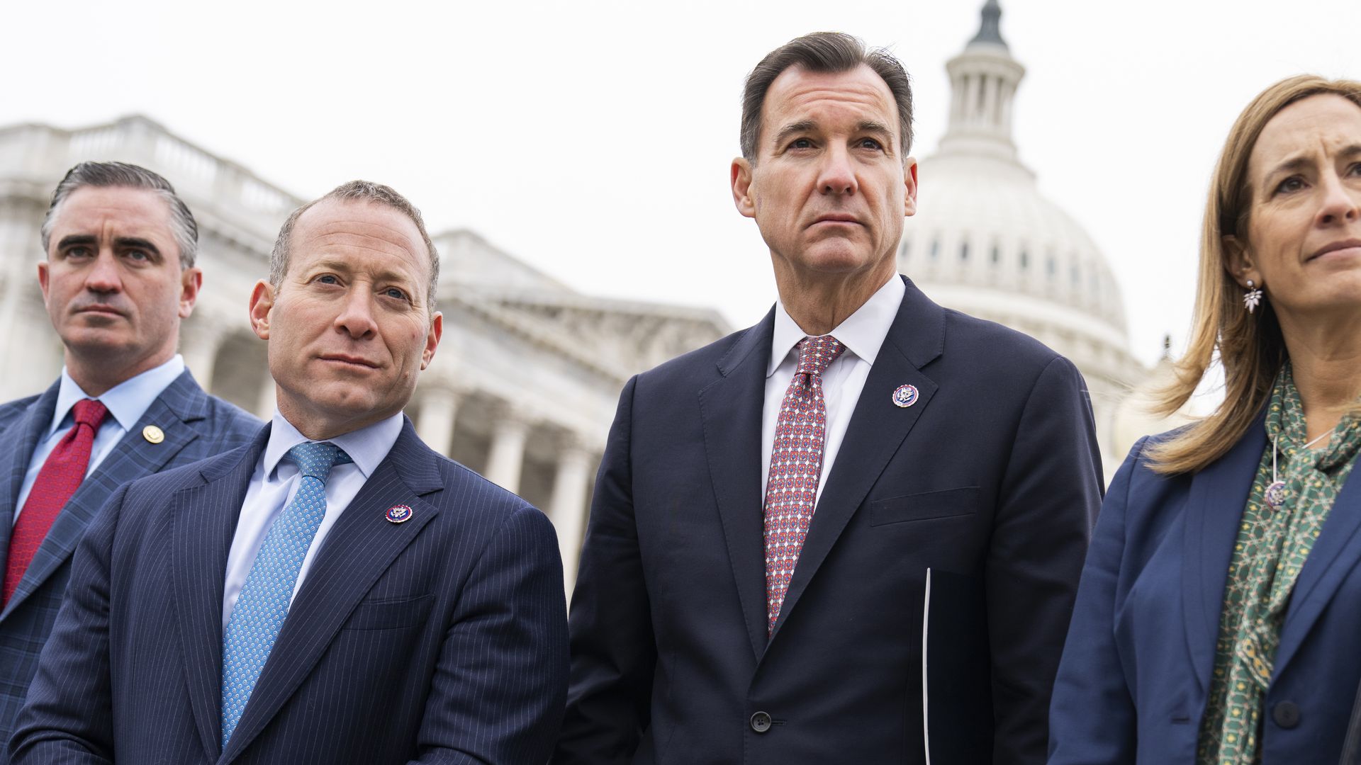 Reps. Gottheimer and Suozzi, wearing blue suits, stand in front of the U.S. Capitol and a hedge.