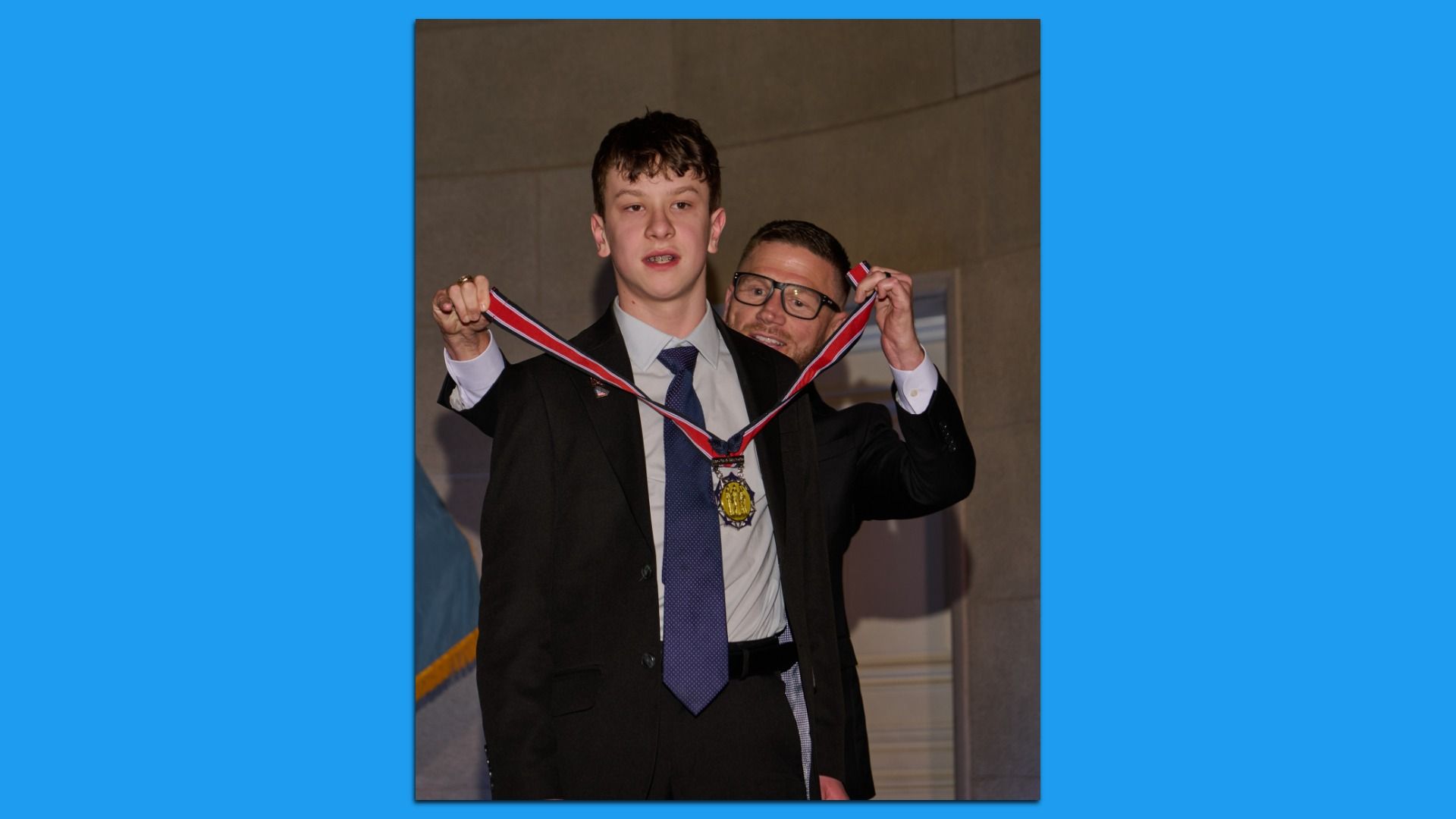 Young man in a dark suit and blue tie receives a gold medal on a red ribbon from an older man with glasses, indoors.