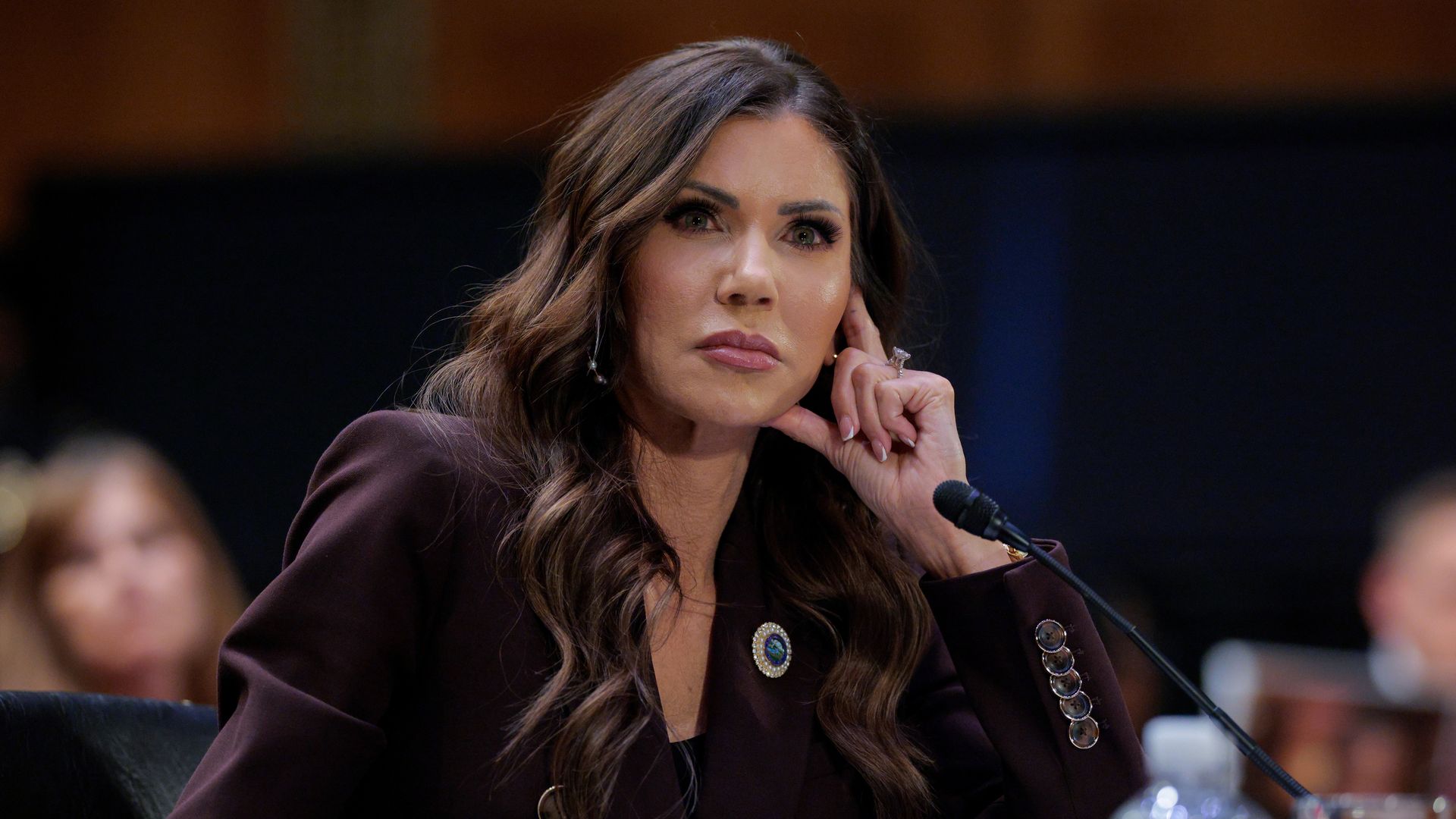 Kristi Noem, wearing a burgundy suit with a blue and silver brooch, leans on her hand as she testifies to lawmakers (not pictured) on Capitol Hill. 