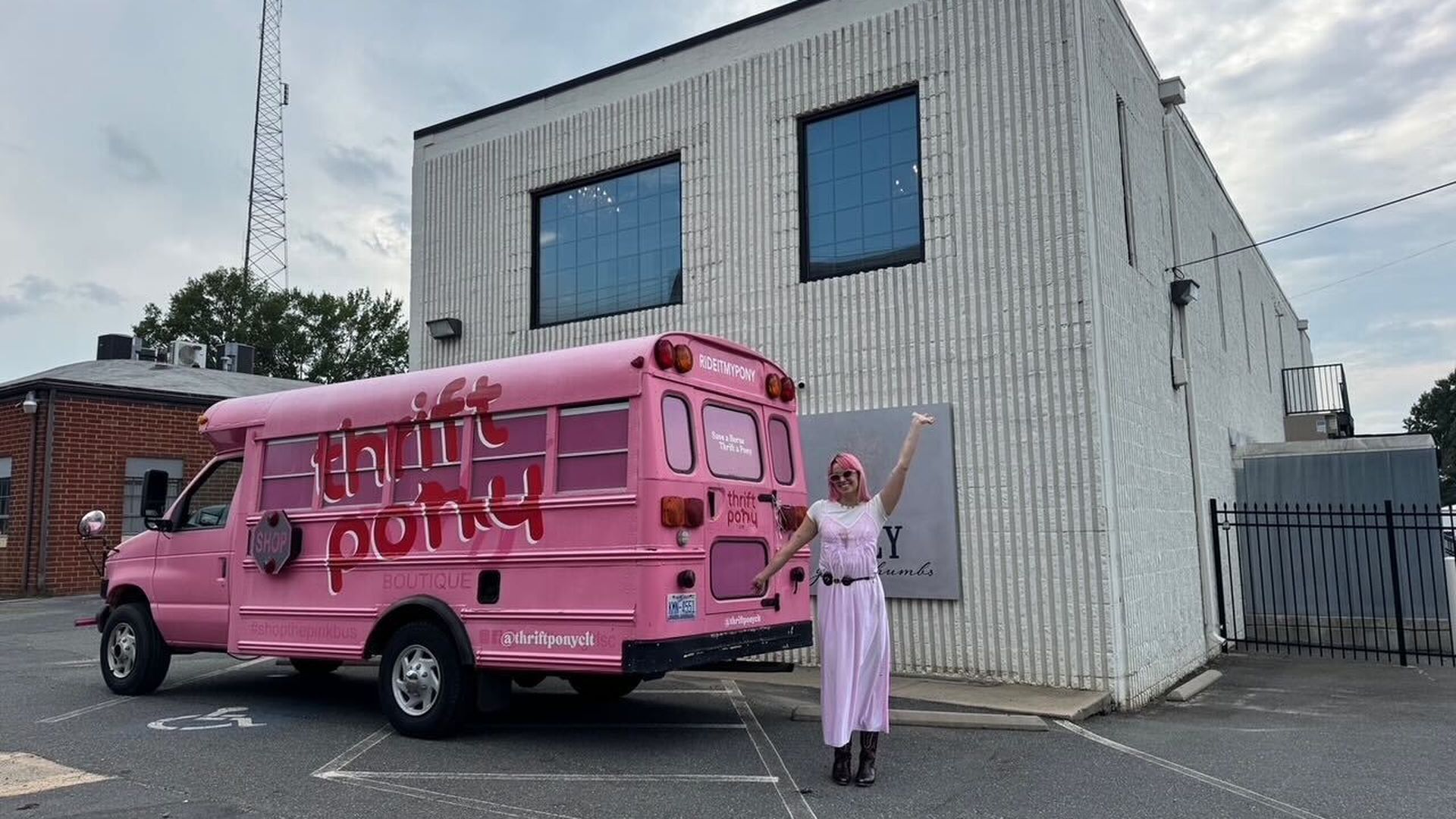 Person dressed in pink and purple stands next to a bright pink bus with "thrift pony boutique" written on its side, parked near a white building under a cloudy sky.