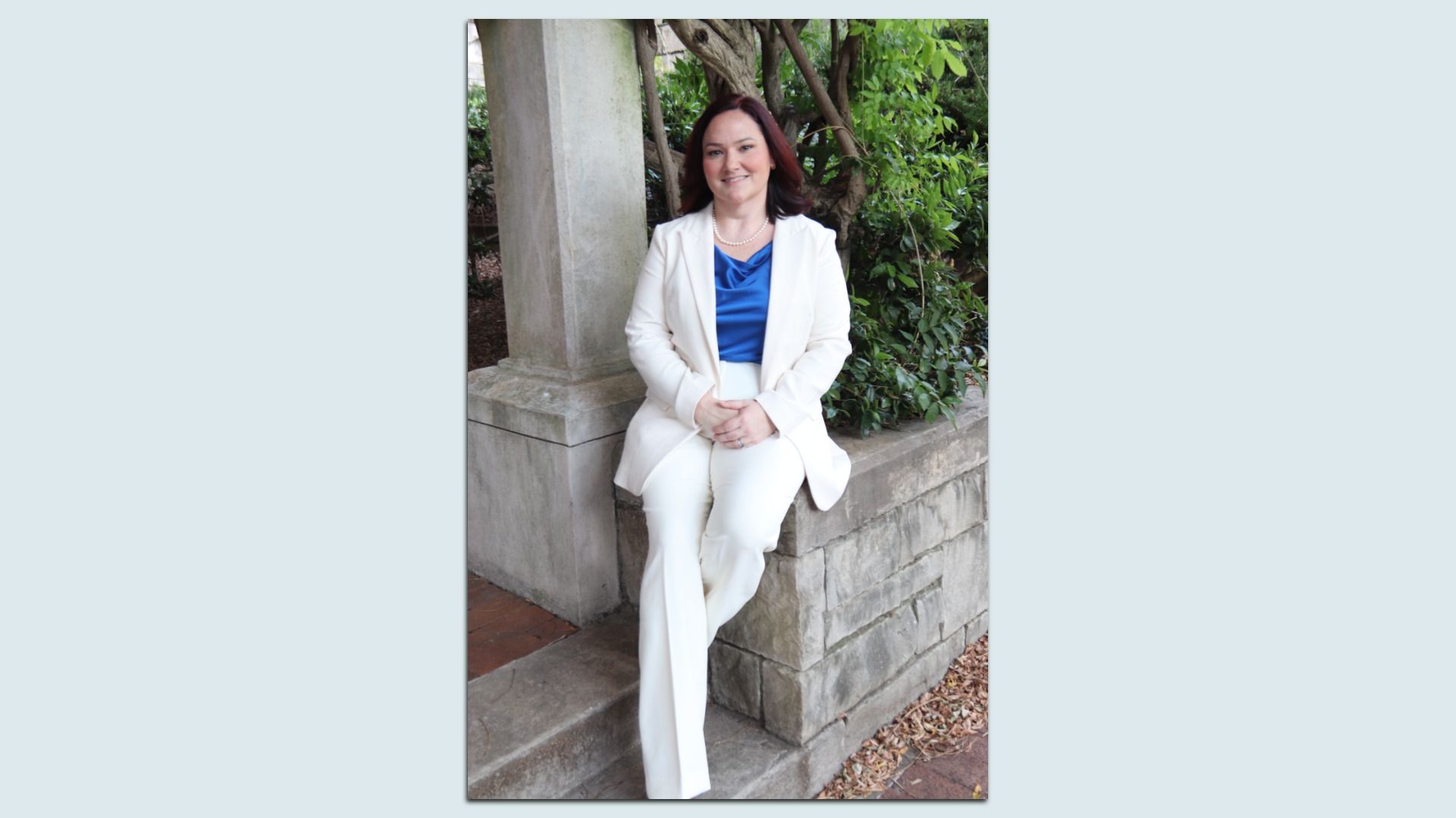 Smiling woman with dark hair sits on a stone ledge beside a column, wearing a white blazer and white pants with a blue blouse and pearl necklace, surrounded by greenery in a garden.
