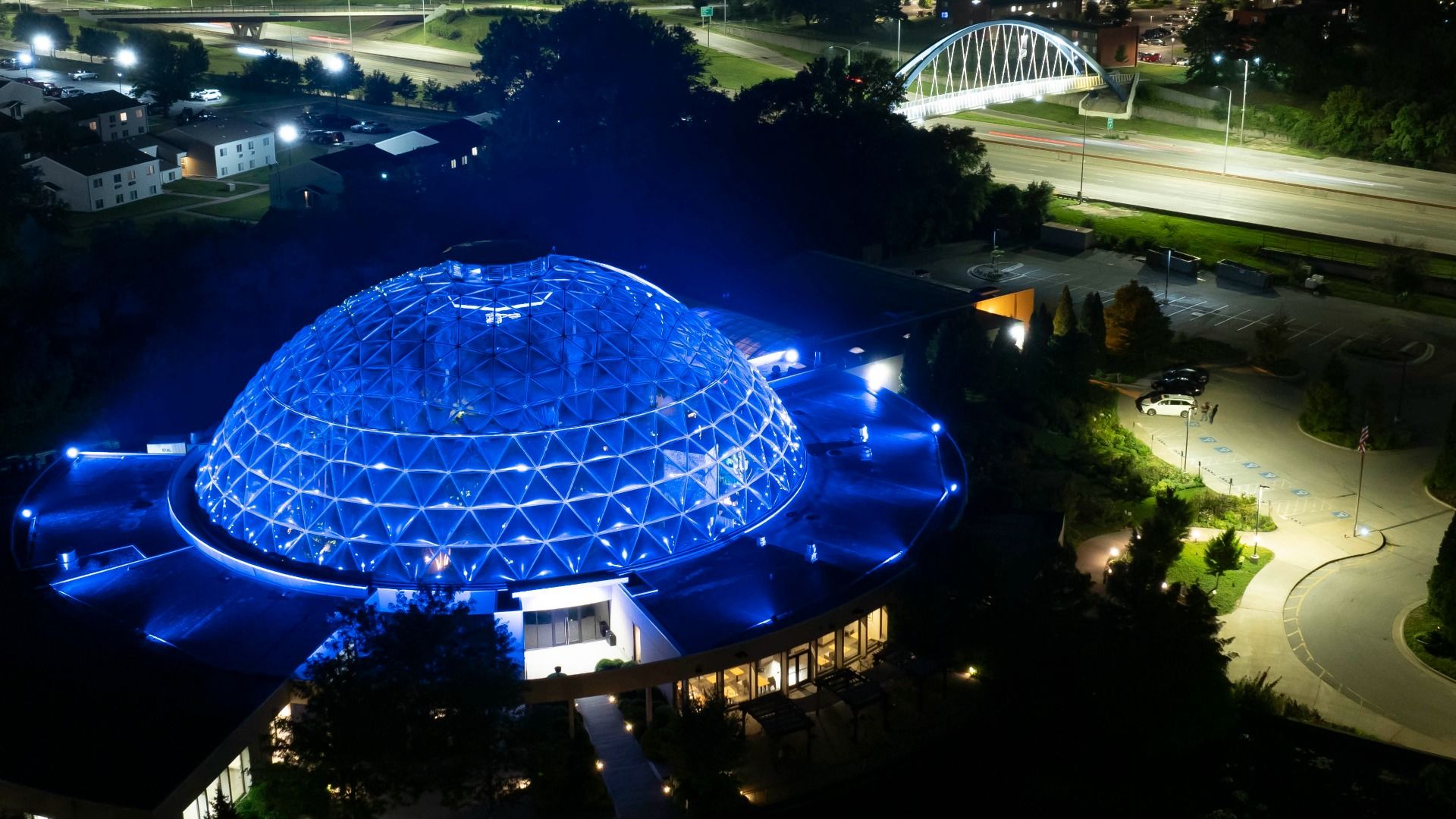 Aerial night view of a large blue illuminated geodesic dome beside a parking lot, with a white arched bridge across a road in the background; tall trees frame the scene.