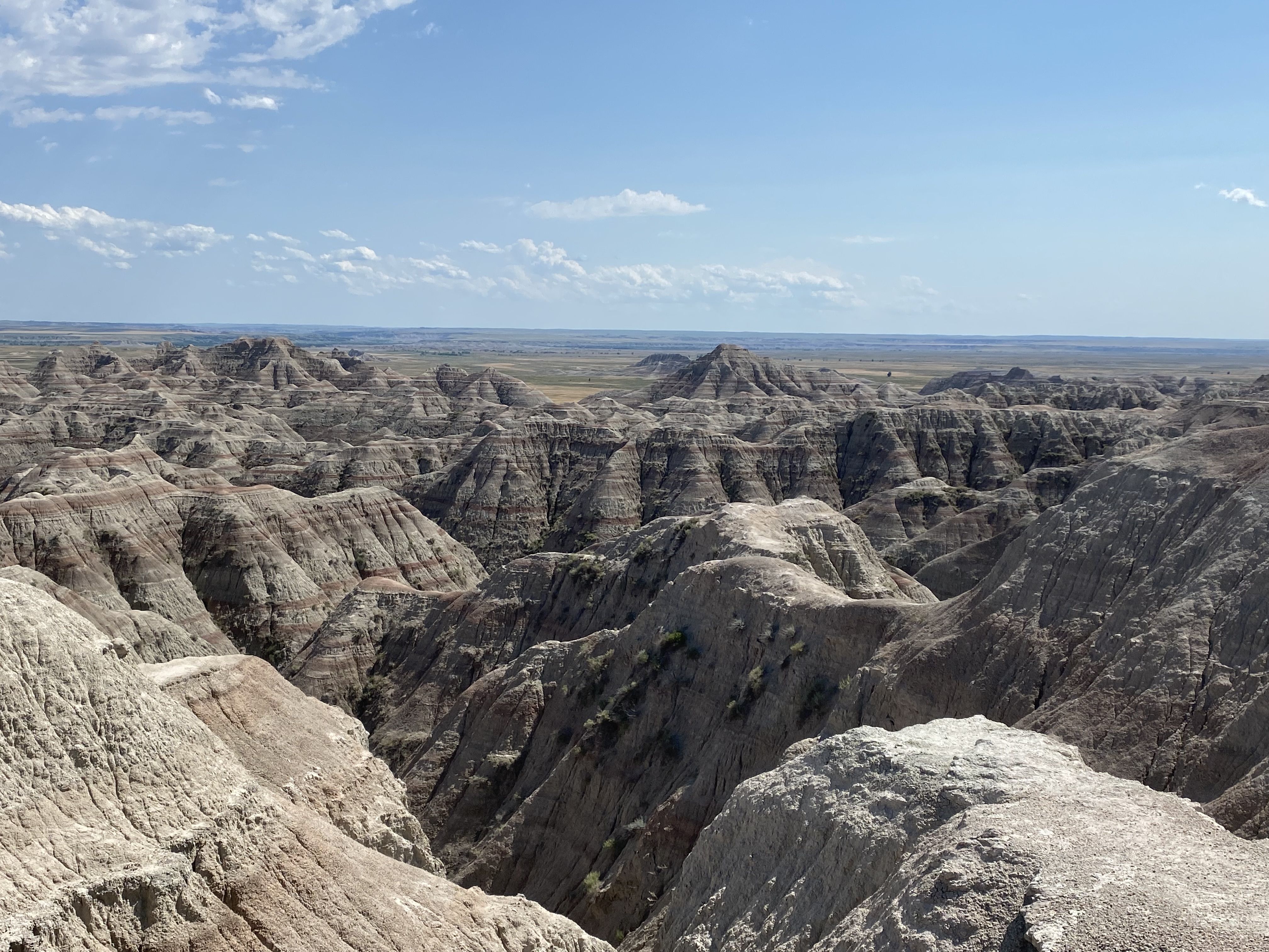 Vast desert badlands with layered, eroded rock formations in the foreground, a flat valley beyond, and a clear blue sky with scattered white clouds.